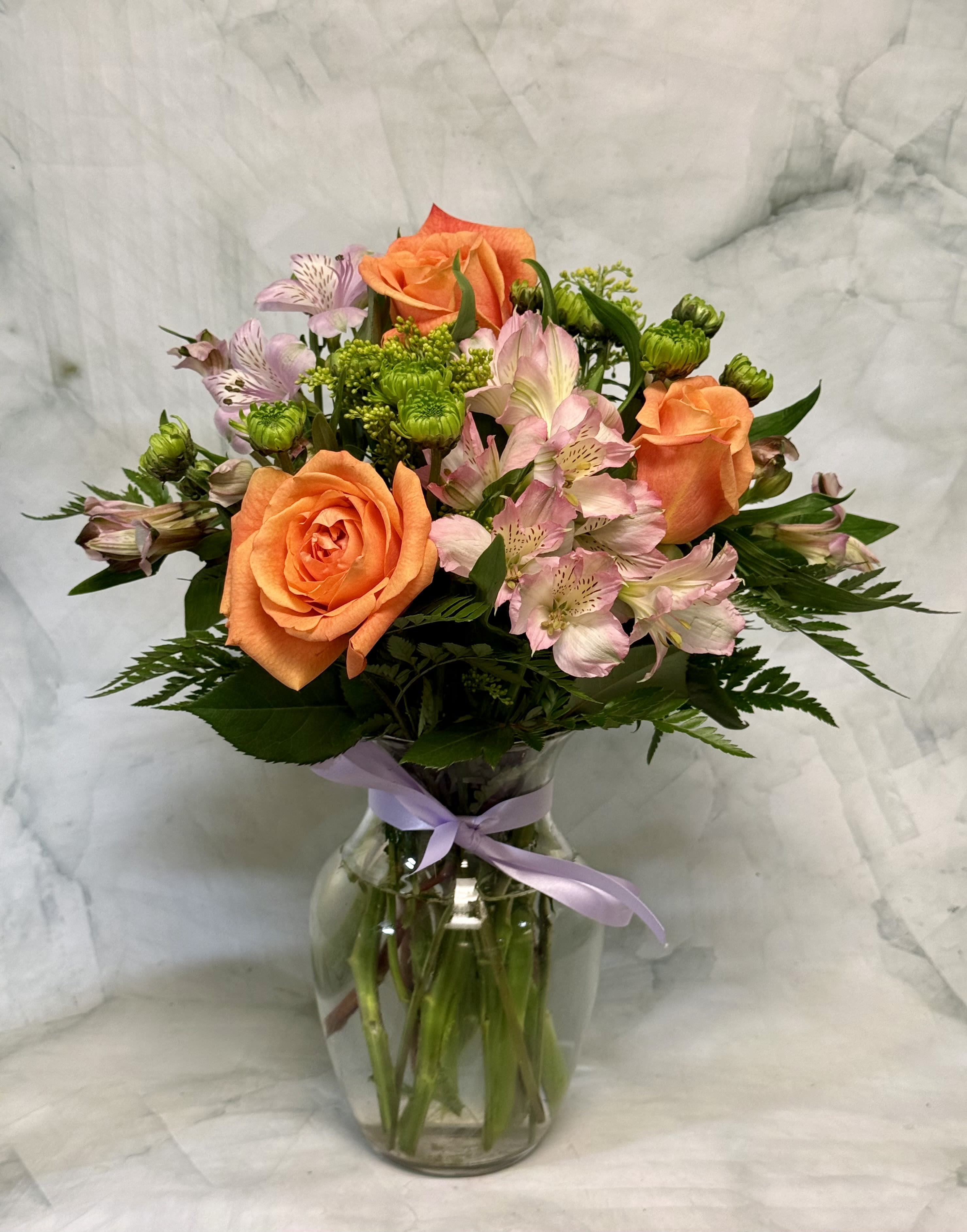 Sunkist - Bright orange roses take center stage in this birthday flower arrangement, their sunlit petals paired with soft pink alstroemeria for an uplifting, warm feeling that says "thinking of you." Perfect as a birthday bouquet or a bright get-well pick-me-up, Sunkist also reads as a sincere thank-you or gentle I'm-sorry gesture-ideal for spring and summer moments when fresh, cheerful color matters.  This arrangement mixes orange roses, pink alstroemeria, and small green chrysanthemum blooms with complementary white accents for a balanced, rounded silhouette. Flowers arrive arranged in a clear glass vase tied with a lavender ribbon, giving the design a simple, polished look that works on a bedside table or dining-room centerpiece. The combination of long-lasting varieties means the recipient will enjoy blooms for days.  Send this when you want someone to feel genuinely seen and cheered-the bright hues and airy textures make it the kind of bouquet that brings immediate warmth. Available in a classic vase size; care is simple: fresh water and a cool spot will help extend bloom life.