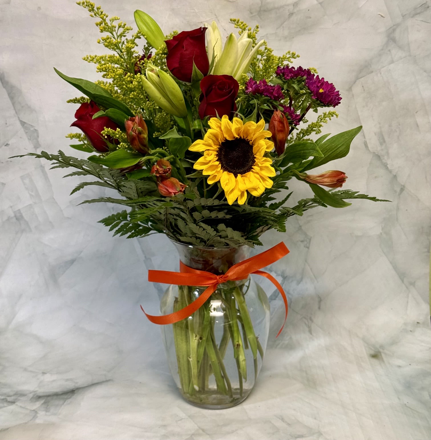 Summer Breeze - A bright sunflower crowns this Summer Breeze flower arrangement, making it an uplifting choice for a summer gift or a Father's Day bouquet. The centerpiece sunflower and deep red roses create a warm, celebratory contrast that instantly reads as joyful and thoughtful for birthdays, thank-you gestures, or just because you're thinking of them.  This full, upright arrangement mixes pale lily buds, chrysanthemums, golden mums, orange alstroemeria accents, carnations, alstroemeria, daisylike dianthus, and delicate roses in a clear glass vase tied with a vivid orange ribbon. The colors-sunny yellow, rich red, soft white, and pops orange-play together for a balanced, textured look. The vase presentation arrives ready to display and makes the bouquet easy to place on a desk, kitchen counter, or bedside table.  Made to make the recipient feel genuinely celebrated, Summer Breeze suits casual summer gatherings as well as special moments like Father's Day. Standard care-fresh water and a cool spot out of direct sun-will help the mixed blooms last longer; the compact clear vase keeps stems supported for an attractive, long-lasting gift.