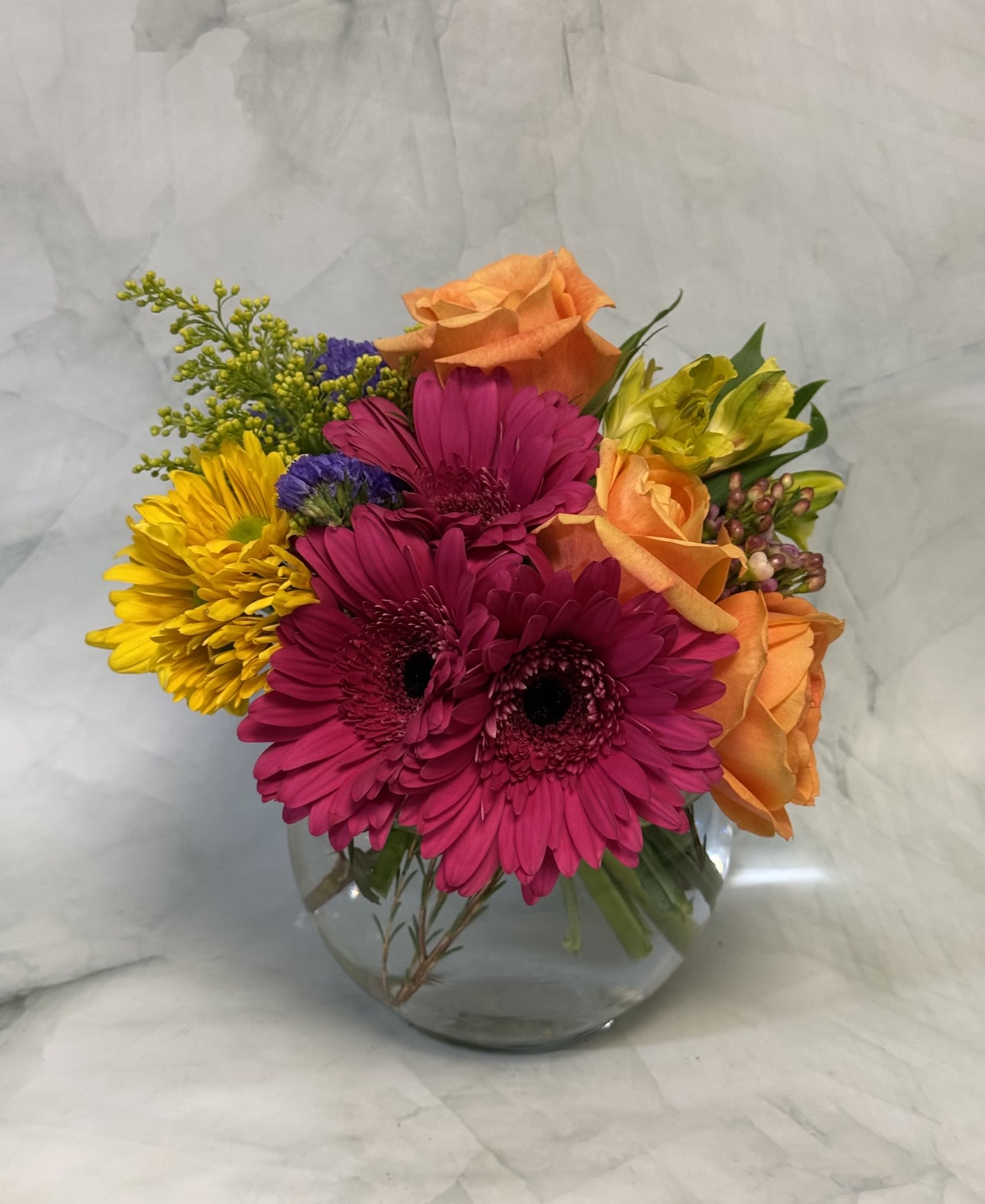 Sophisticated Summer - Bright magenta gerbera daisies and warm orange roses give this flower arrangement a lively, celebratory pulse-perfect as a summer bouquet or a thoughtful graduation gift. A mix of yellow blooms, pink and purple accents, and green sprays creates a vivid, upbeat palette that reads as both joyful and refined, an ideal way to say "thank you" or "congrats" to the person you're thinking of.  Arranged in a low, rounded clear glass vase, the bouquet combines alstroemeria, asters, chrysanthemums, daisies, gerbera daisies, roses, and delicate purple filler for contrast and texture. The compact, dome-like shape keeps the colors tightly grouped so the magenta and orange focal flowers pop against softer yellow and pink tones. Approximate vase style and size: clear glass low vase suitable for tabletops and mantels.  This is the kind of mixed bouquet that makes someone feel truly celebrated-bright, cheerful, and effortless to display. It's ready to place in the vase provided; for best longevity, keep in fresh water and out of direct heat.