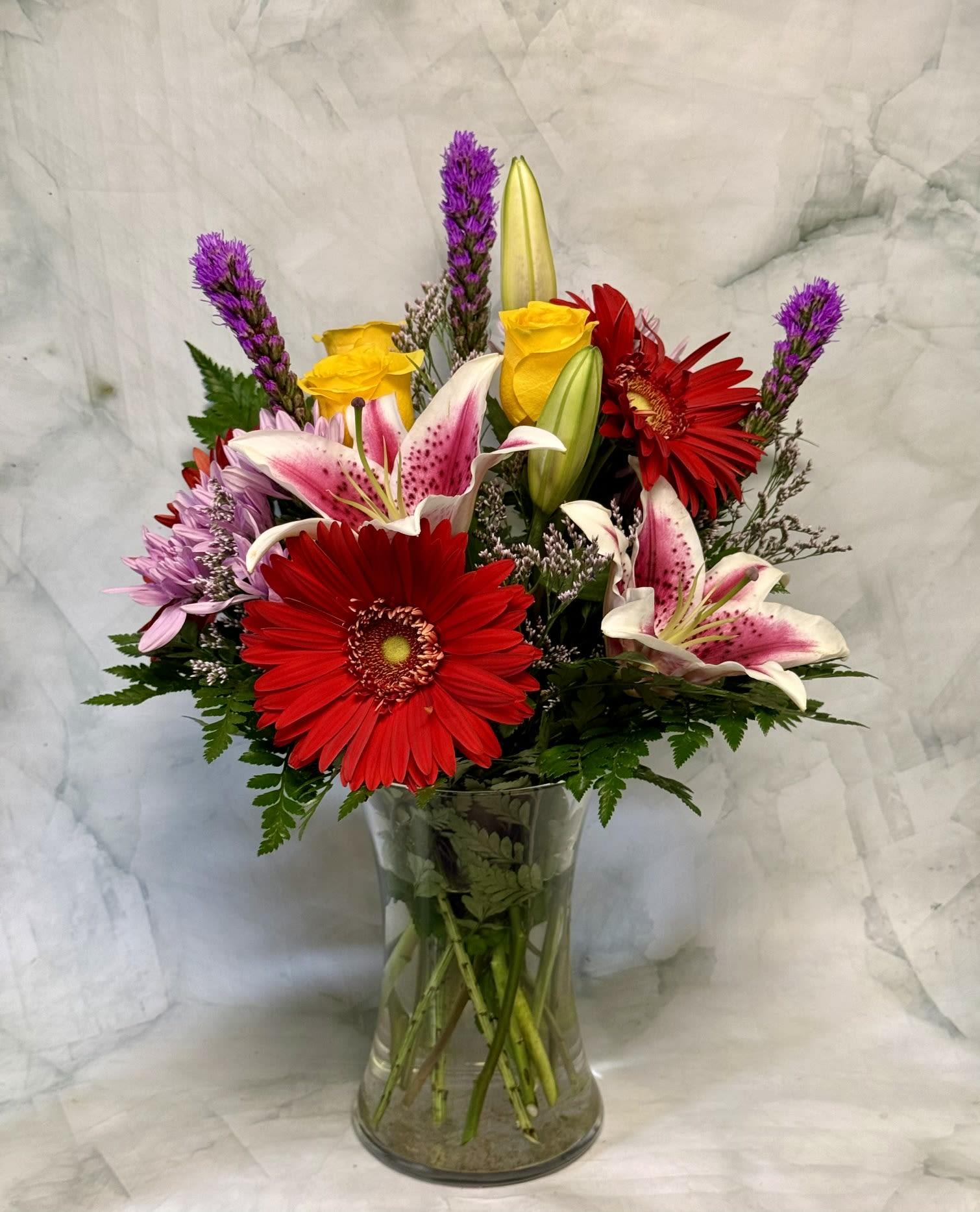 Summer Lovin' - A bold orange gerbera takes center stage in this Summer Lovin' flower arrangement, paired with pink-and-white lilies, sunny yellow roses, and soft pale aster blooms for a bright, cheerful birthday bouquet or just-because gift. The warm, summery palette reads joyful and fresh-the kind of arrangement that makes the person you're thinking of feel noticed and celebrated.  Flowers are gathered in a clear glass vase with dark foliage at the base and a few purple accent spikes rising above the main cluster for height and contrast. Expect a lively mix of chrysanthemums, gerbera daisies, lilies, roses, and asters in orange, pink, yellow, white, and complementary accents; the vase presentation is ready to display on a table or desk.  Perfect for summer entertaining, a thank-you, or sending a quick pick-me-up, this arrangement arrives arranged and trimmed for immediate enjoyment-simply place in a cool spot out of direct midday sun and refresh the water every few days to prolong bloom life.