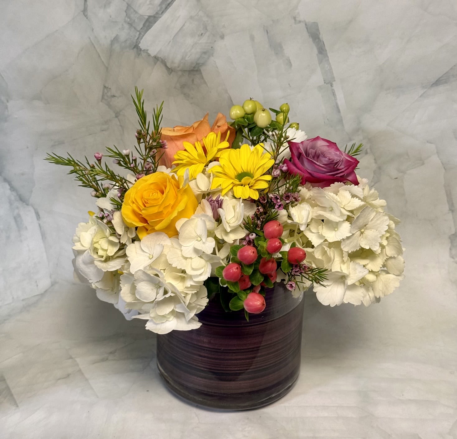 Springtime Magic  - A lively burst of spring in a ceramic vase, this flower arrangement is perfect as a Get Well, Just Because, or Thank You gift that will brighten someone's day. The mix of white hydrangeas, pink and peach roses, and cheerful yellow daisies reads fresh and optimistic-an ideal pick for spring, Passover, or Easter visits while still feeling right any time you want to lift someone's spirits.  Carefully balanced for color and texture, the design pairs large, rounded hydrangea heads with a magenta rose, a soft peach rose, a sunny yellow rose, and bright yellow daisy blooms. Small pink berry-like accents and fine-textured filler weave between the focal flowers, adding delicate contrast against the fuller petals. The arrangement sits in a dark ceramic cylinder vase that gives the composition a contemporary, grounded look and makes it easy to display on a bedside table, entryway console, or kitchen counter.  Made to make the recipient feel seen and cared for, this bouquet says "thinking of you" without fuss. Choose a size that fits the space-compact for a bedside pick-me-up or larger for a centerpiece-and refresh the water every few days to keep blooms looking their best.