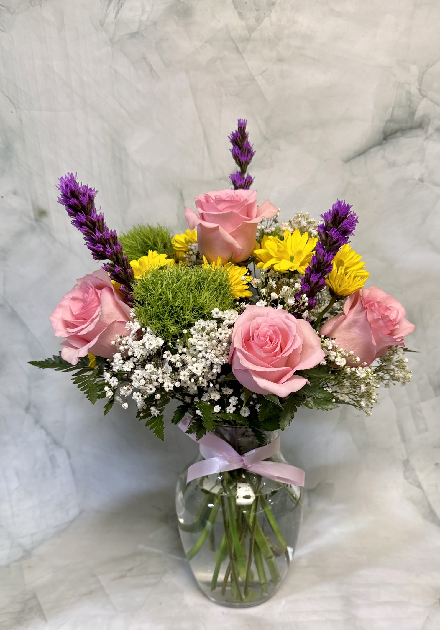 Colorful Spring Meadow - Soft pink roses and bright yellow daisy-like chrysanthemums meet airy white asters and playful green textural blooms in a bouquet that reads like a Colorful Spring Meadow - perfect as a birthday bouquet, a cheerful Admin Professionals' Week gift, or a fresh "congrats" surprise. The tall purple spikes add dramatic height and a whimsical contrast that makes the recipient feel genuinely celebrated and noticed.  Arranged in a clear glass vase tied with a soft pink ribbon, this garden-style arrangement pairs rounded green elements and delicate dianthus with mixed filler flowers for a layered, textured look. Colors include pink, yellow, green, purple, and white; stems are styled to create both height and fullness so it photographs beautifully and fills a tabletop or desk.  Designed for spring but lovely any time someone needs brightening, this bouquet is an easy way to say thank you, well done, or thinking of you. Vase included; for best longevity, refresh the water every 2-3 days and trim stems slightly when you notice drooping to keep blooms perky.