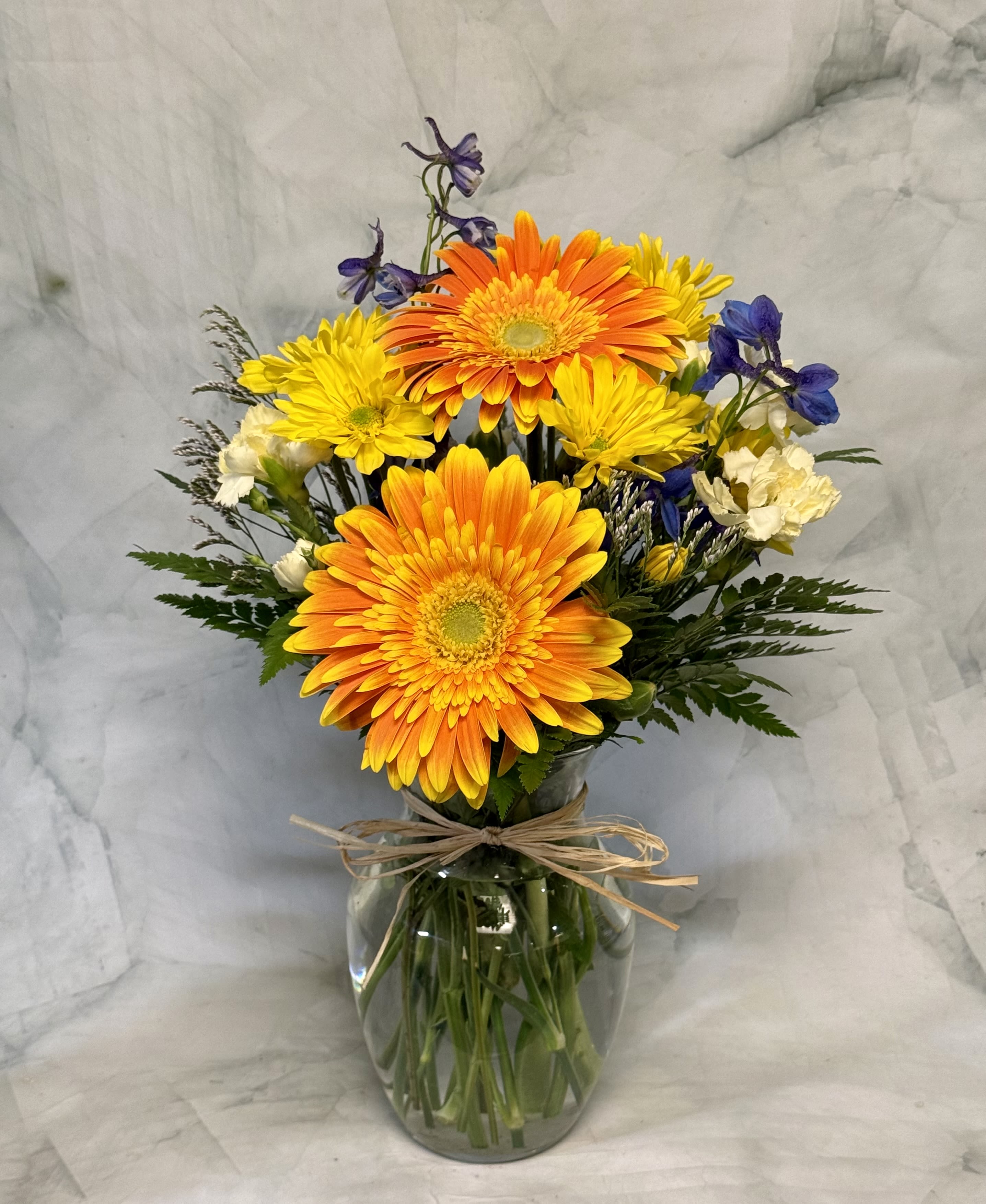 Life is a Beach!  - Two large gerbera daisies-sunny yellow or vibrant orange depending on the selection-give this summer flower arrangement its instant, beachy charm, making it a perfect graduation bouquet or a bright Fourth of July gift. The casual hand-tied look in a clear glass vase with a raffia bow reads like a relaxed seaside postcard, designed to make the person you're thinking of feel genuinely celebrated and seen.  Surrounding the focal gerberas are sky-blue delphinium spikes, pale green mini carnations, lavender accents, yellow button poms and touches of white and deep green foliage for contrast. The loose, airy silhouette and mix of bold yellow, cool blue, soft purple and fresh green create a breezy, upbeat palette that works for birthdays, thank-you gestures, or just-because moments. Presented in a standard clear glass vase tied with raffia, it's easy to place on a table or bring along to a casual get-together.  Care is simple: fresh water and a cool spot out of direct sun will keep the blooms looking their best. Sized for easy gifting, this arrangement reads year-round as a sunny, relaxed pick-me-up-especially welcome during summer celebrations and milestone graduations.
