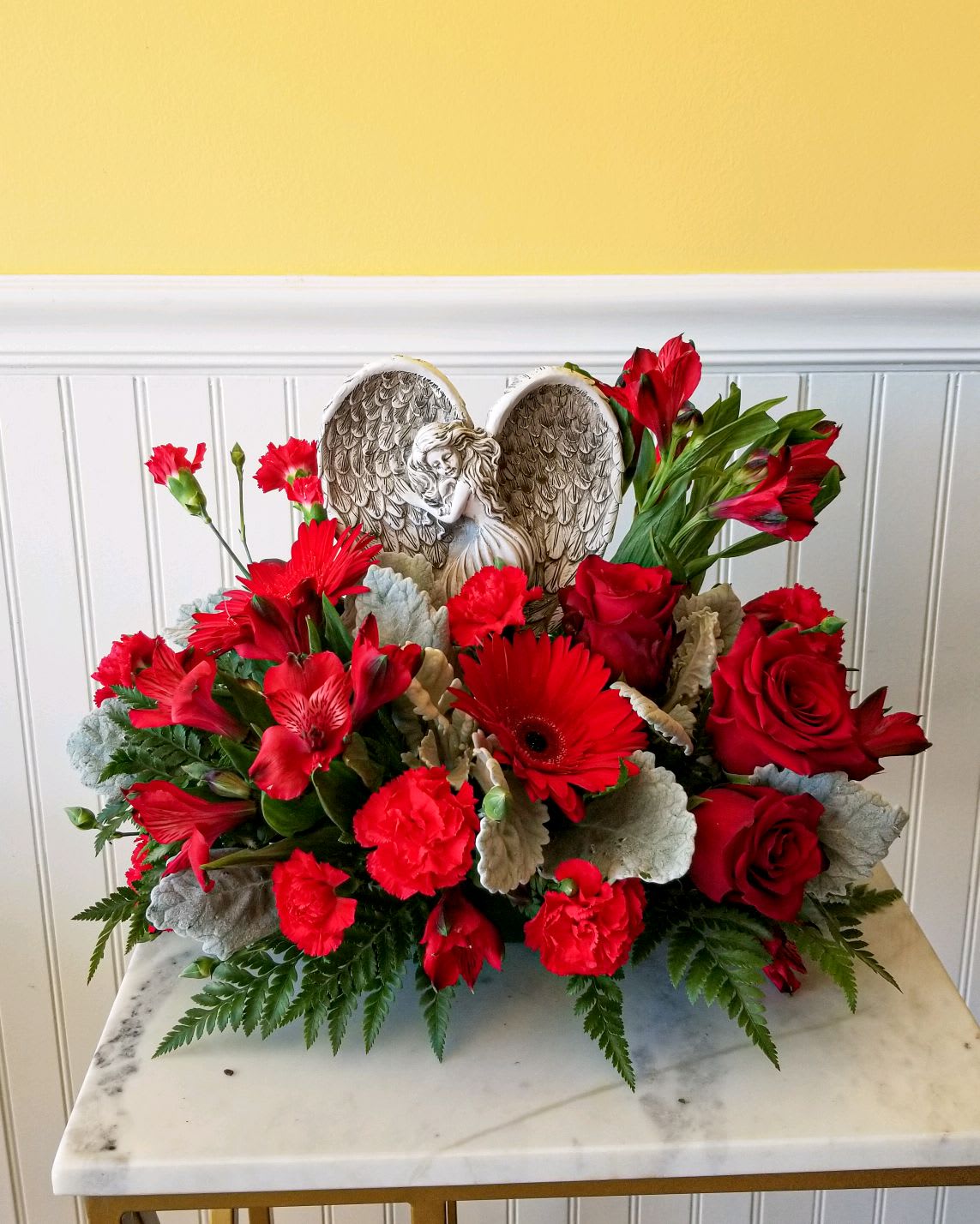 Red floral arrangement with roses, gerbera daisies, and carnations around an angel wing ornament