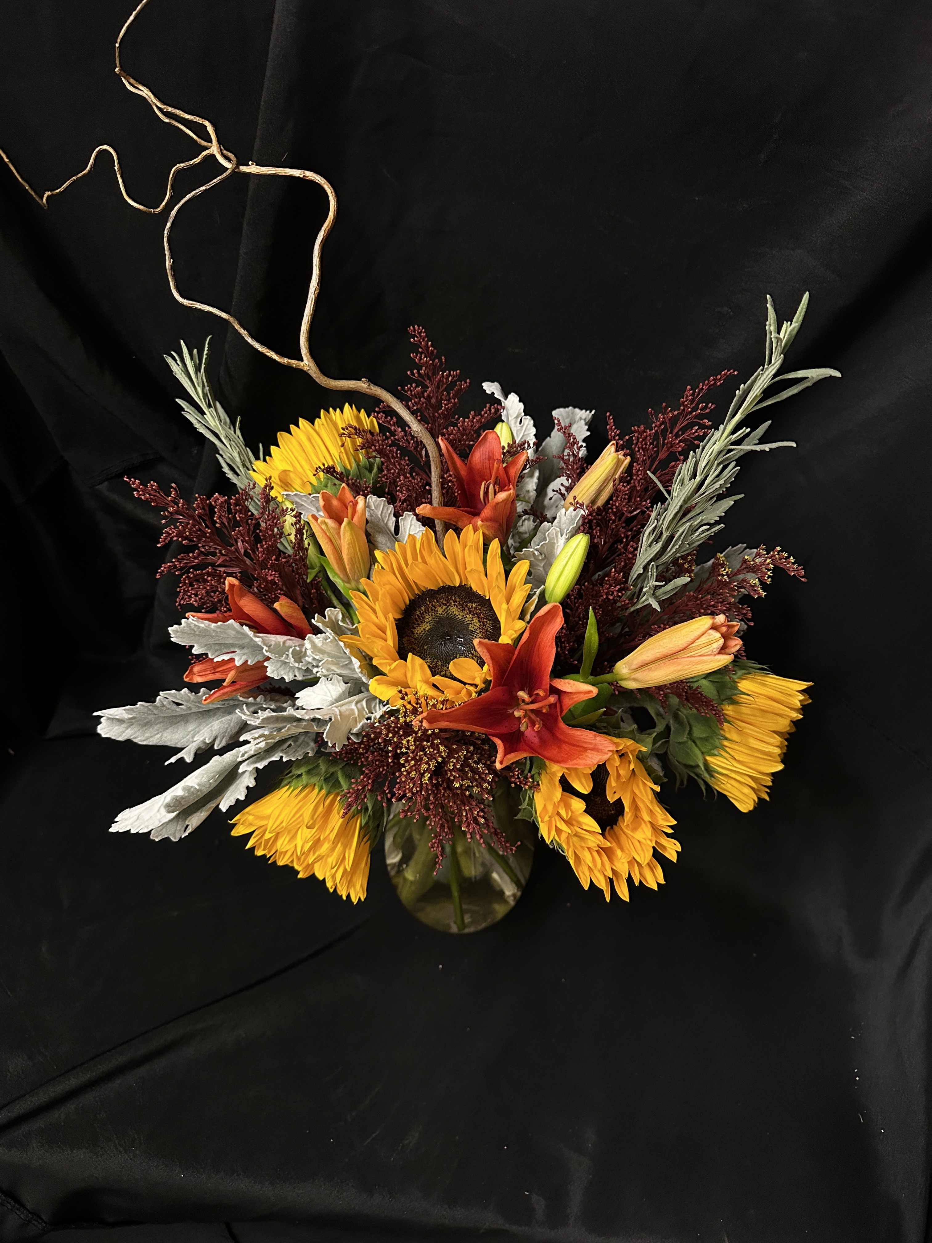 Sunflowers and orange lilies arranged in a glass vase with curly branches