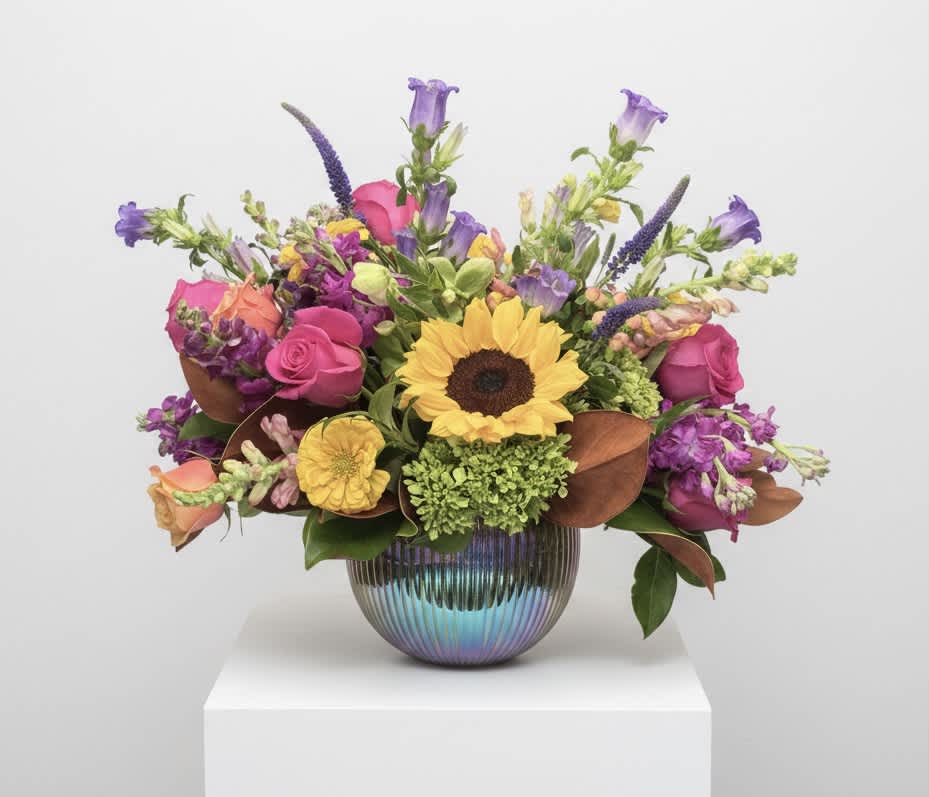 Mixed bouquet with a sunflower, pink roses, and purple blooms in a ribbed iridescent vase