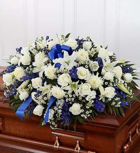 White and blue funeral casket spray with roses, lilies, and ribbon on a wooden casket