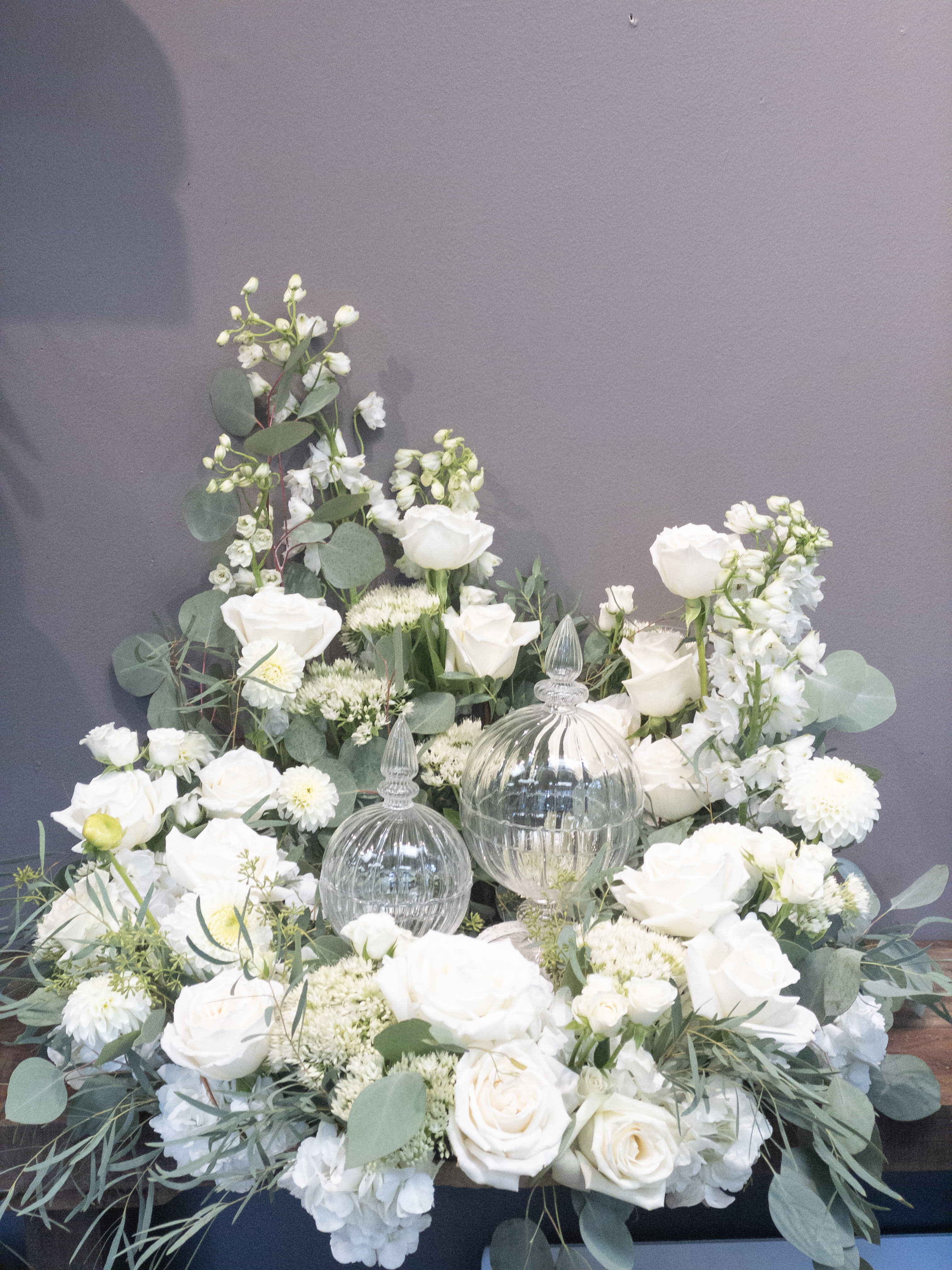 Low white flower arrangement with roses and mums surrounding two clear glass lidded jars.