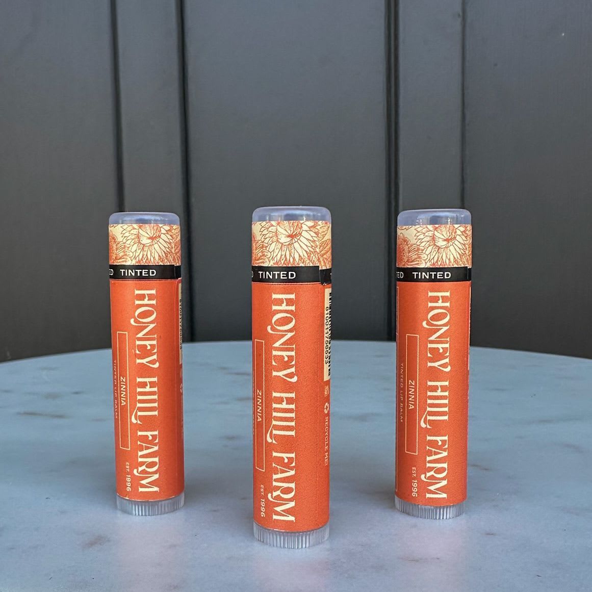 Three orange Honey Hill Farm lip balm tubes standing on a round light blue surface