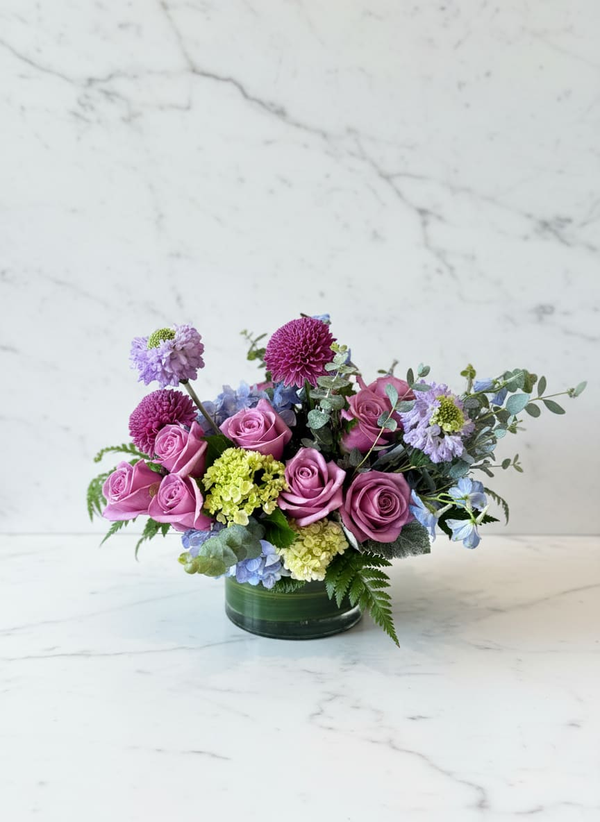 Low arrangement of mauve roses with purple mums and green and blue hydrangea in a glass vase
