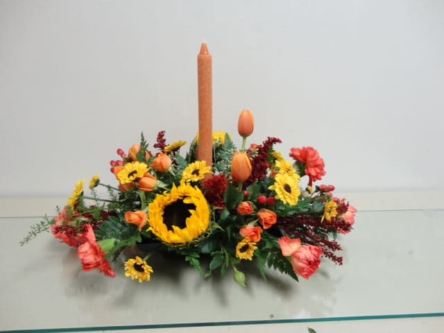 Low floral centerpiece with a tall orange candle and mixed orange, yellow, and red blooms