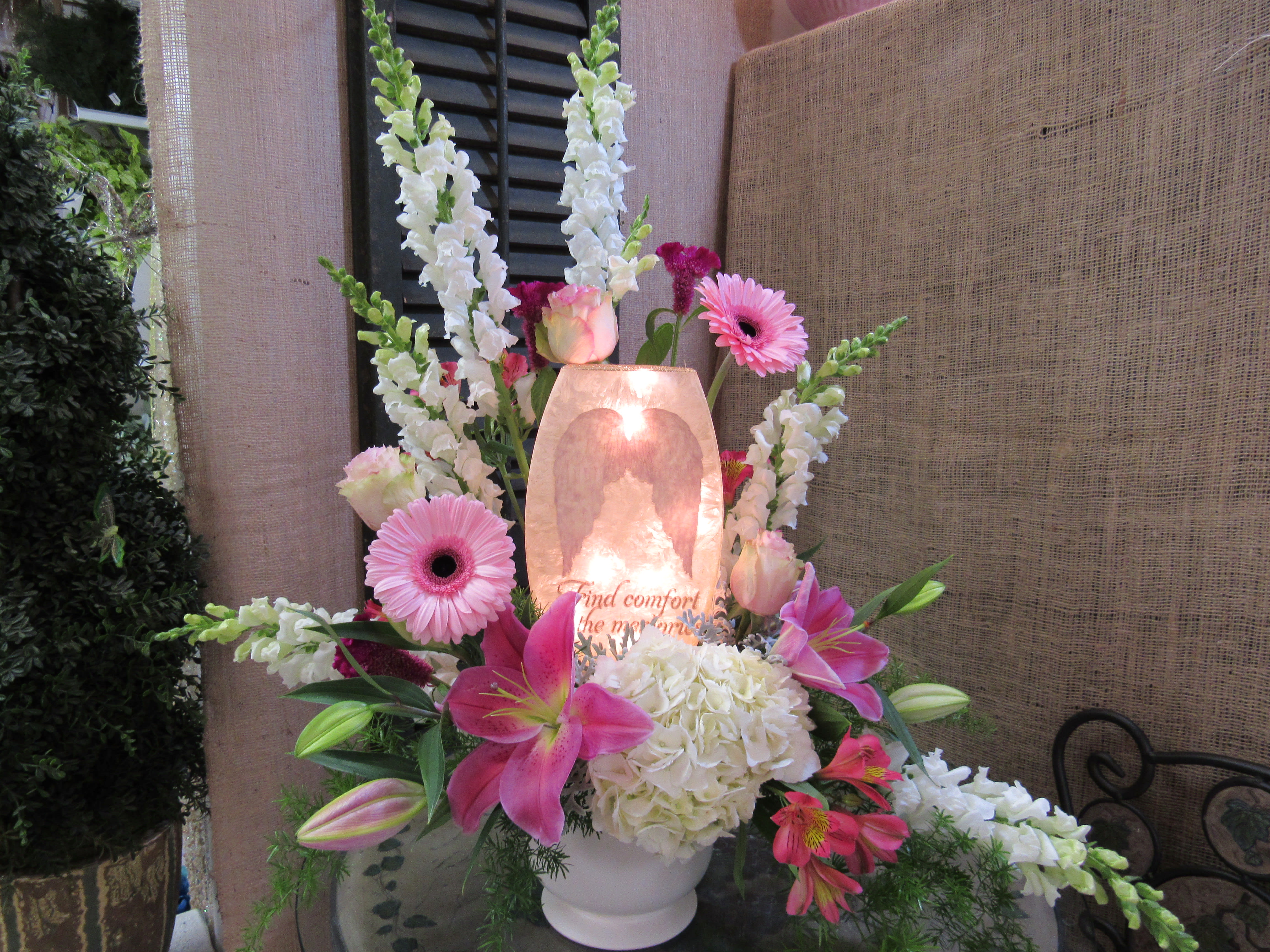 Pink lilies and gerbera daisies arranged around a glowing memorial candle.