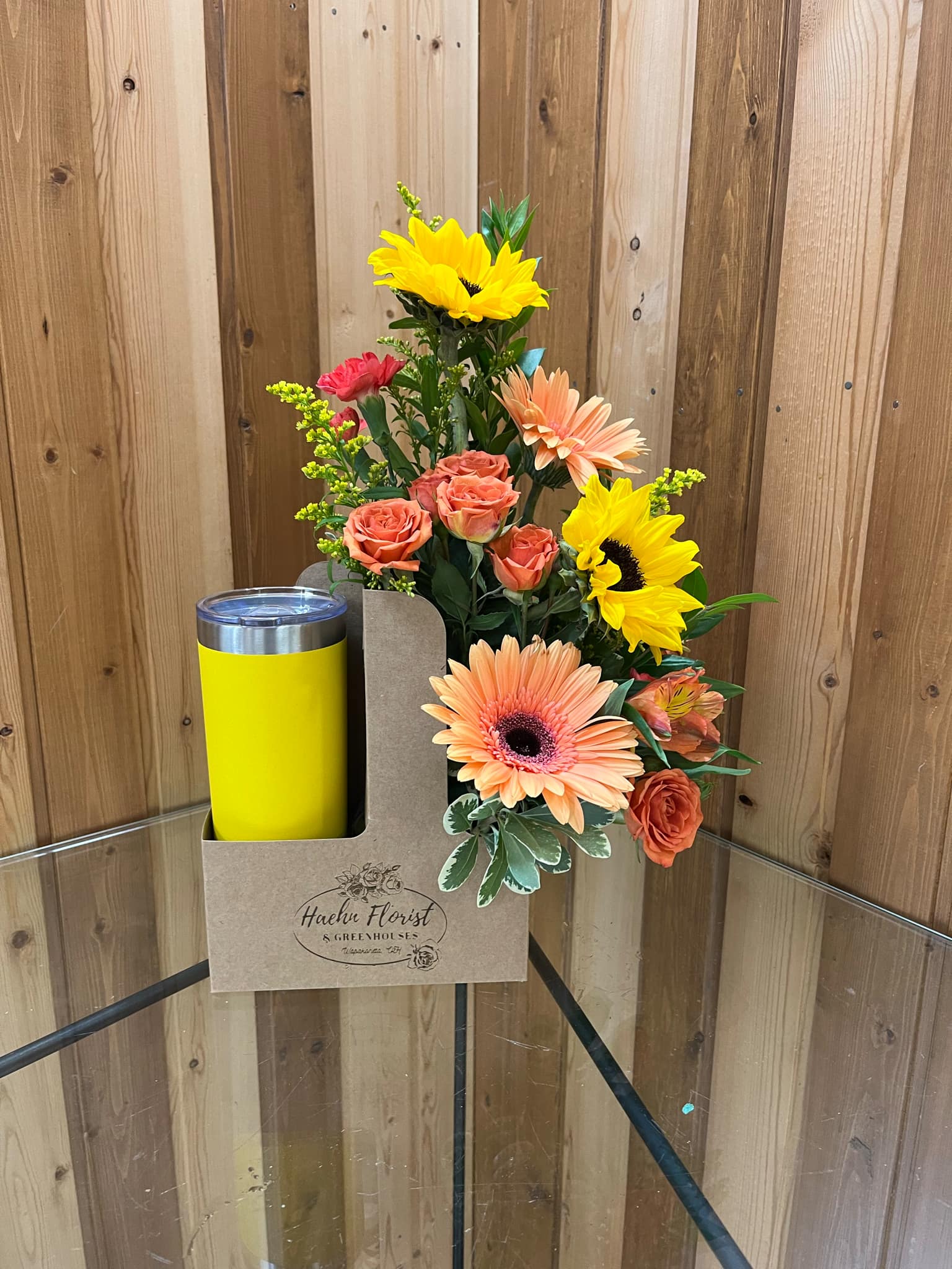 Mixed bouquet with sunflowers, roses, and gerbera daisies in a gift box beside a yellow tumbler