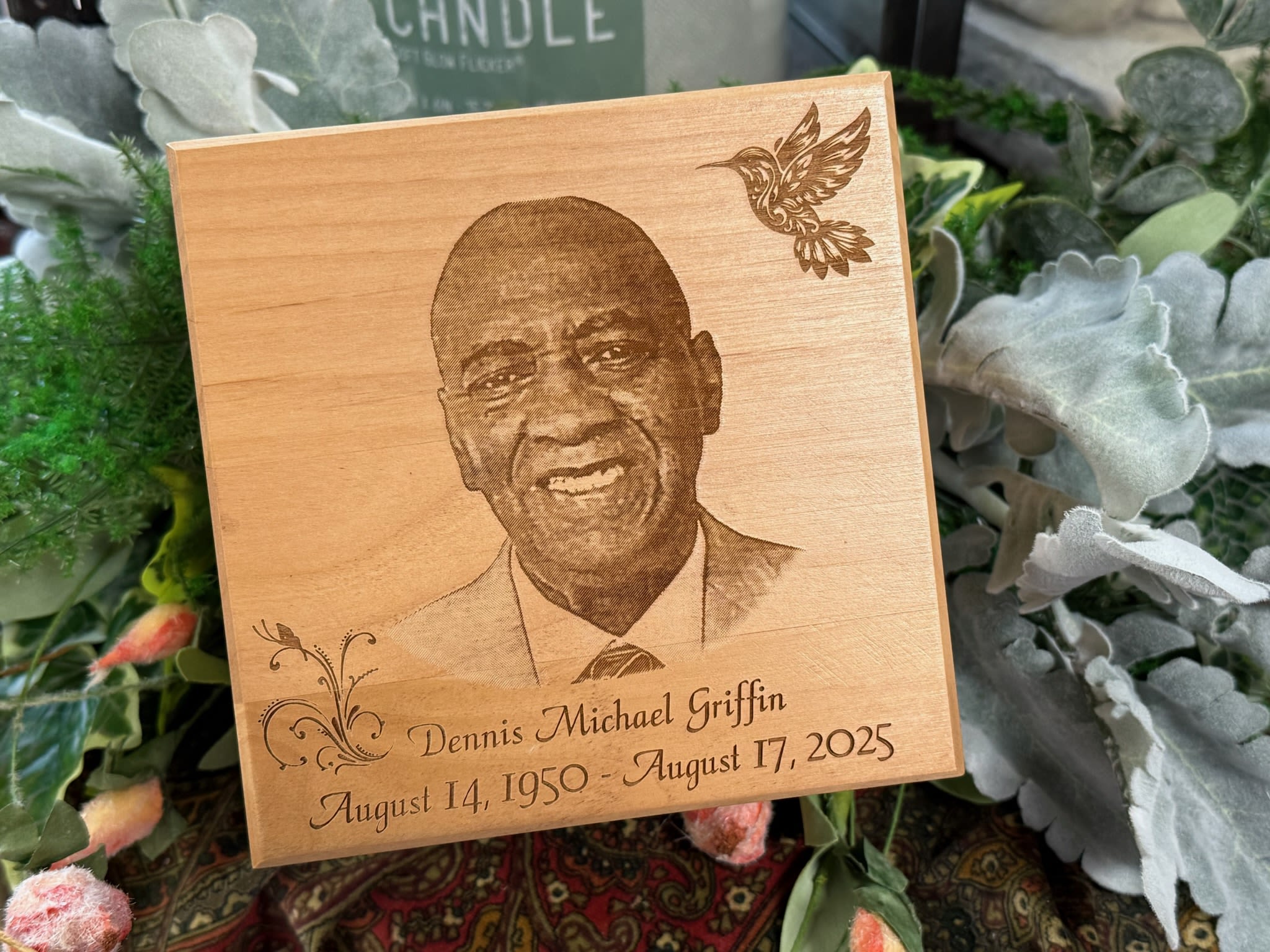 Wooden memorial plaque with a portrait and engraved dates, surrounded by flowers