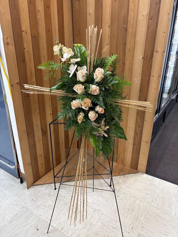Grace &amp; Remembrance Cross Tribute - Honor a cherished life with the timeless beauty of the Grace &amp; Remembrance Cross Tribute. This elegant standing cross features soft blush roses accented with delicate white orchids, lush tropical greenery, and natural bamboo-style accents that create a modern yet peaceful design. The combination of graceful blooms and textured foliage symbolizes faith, hope, and everlasting love, making it a meaningful expression of sympathy and remembrance.  Designed on a standing easel, this striking floral tribute offers comfort to family and friends while celebrating a life filled with love and cherished memories. Ideal for funeral services, memorials, or celebration of life ceremonies.