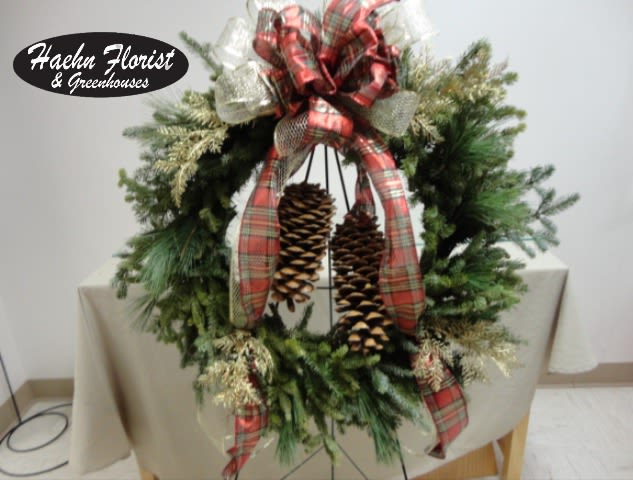 Christmas Fresh Wreath - A classic, full-bodied holiday wreath crafted from fresh mixed evergreens makes a welcoming statement on any front door this winter. This Christmas fresh wreath is built on a sturdy wreath frame and finished with a bold red plaid bow, sheer white mesh ribbon, and metallic accents that pick up the glow of porch lights-perfect for holiday and winter seasonal decorating. The hanging pinecones and natural textures give it a rustic, homespun charm that will make the person you're thinking of feel cozy and celebrated.  Made with dense evergreen branches arranged into a generous circular form, the wreath includes pinecones tucked into the center and wired-in plaid and mesh ribbons for lasting shape and movement. It ships ready to display on a wreath stand or door; approximate diameter and exact stem counts vary with natural material, and substitutions of equivalent fresh evergreens or decorative accents may be made to ensure peak freshness and seasonal availability. Light misting and shelter from prolonged sun or heat will help maintain fragrance and color.  Haehn Florist, Greenhouses, &amp; Flower Delivery - 410 Hamilton Rd., Wapakoneta. For updates and seasonal photos visit www.facebook.com/haehnflorist.