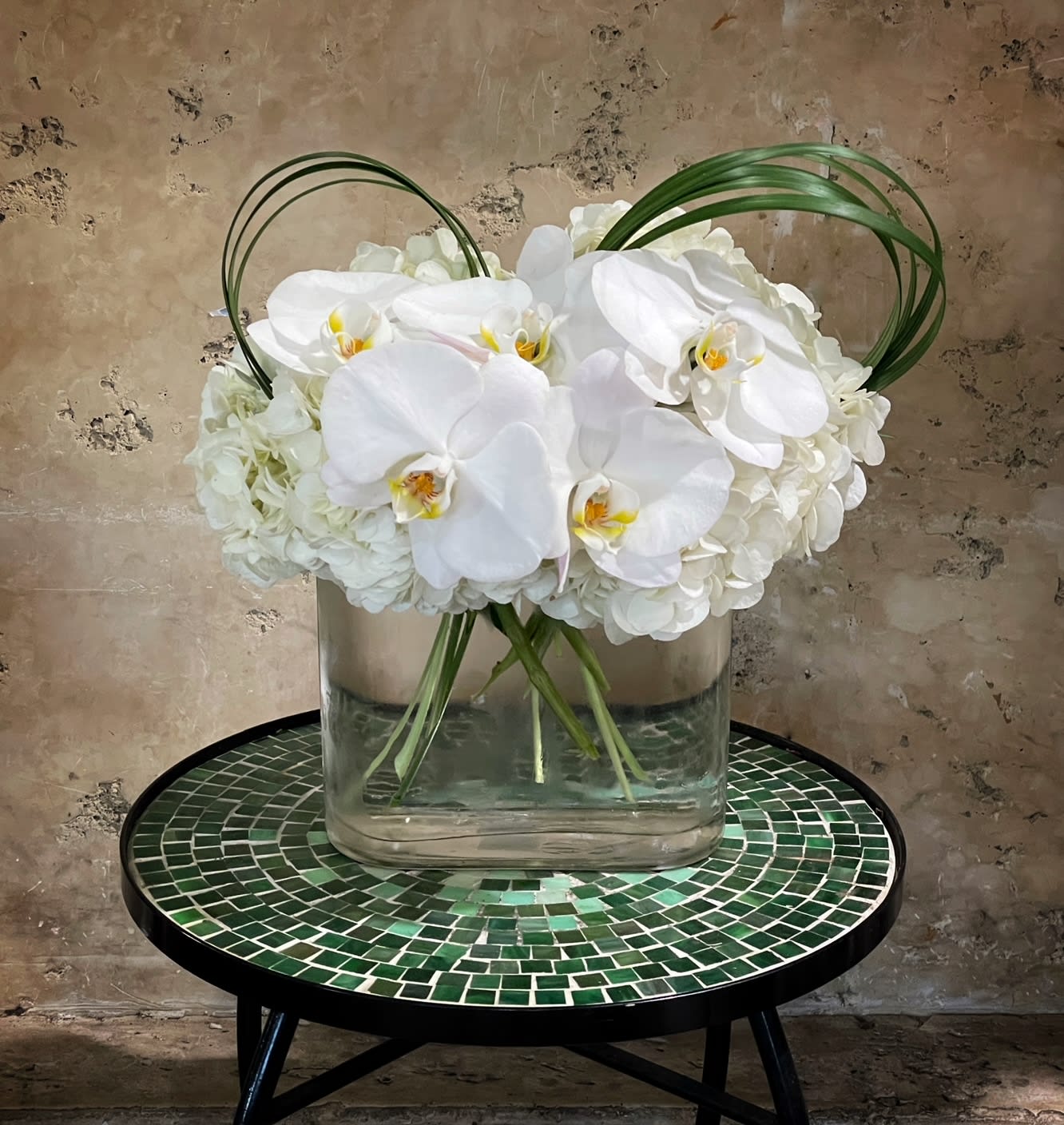 White orchids arranged in a clear glass vase with arched grass loops