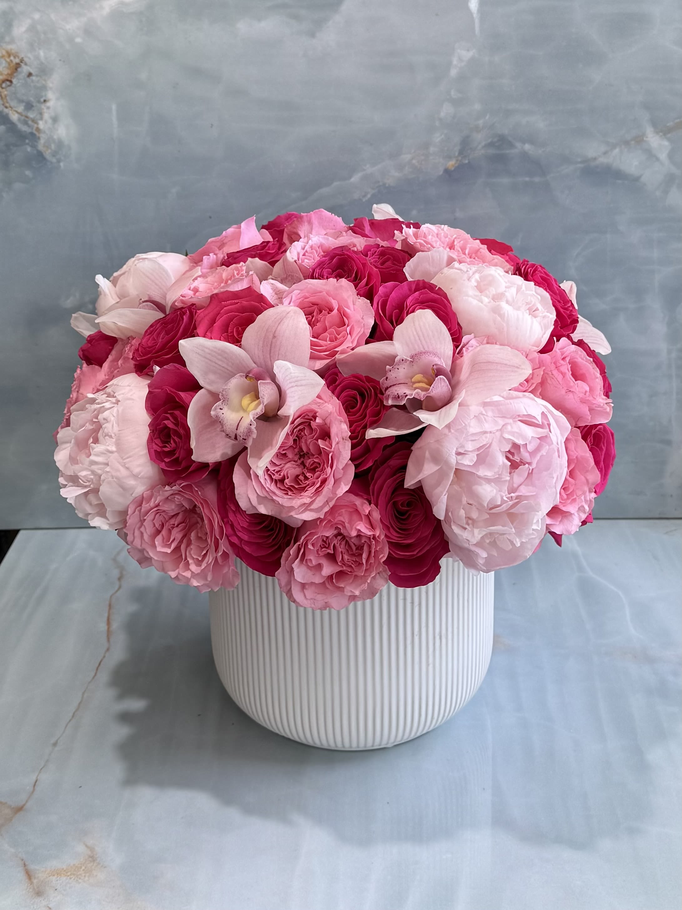 Valentina  - A stunning expression of elegance and romance, this lavish arrangement features layers of blush peonies, premium garden roses, and graceful cymbidium orchids. Designed in a contemporary white ceramic vessel, it offers a beautiful balance of texture, depth, and sophistication-perfect for celebrating life's most memorable moments or bringing everyday luxury into any space. 