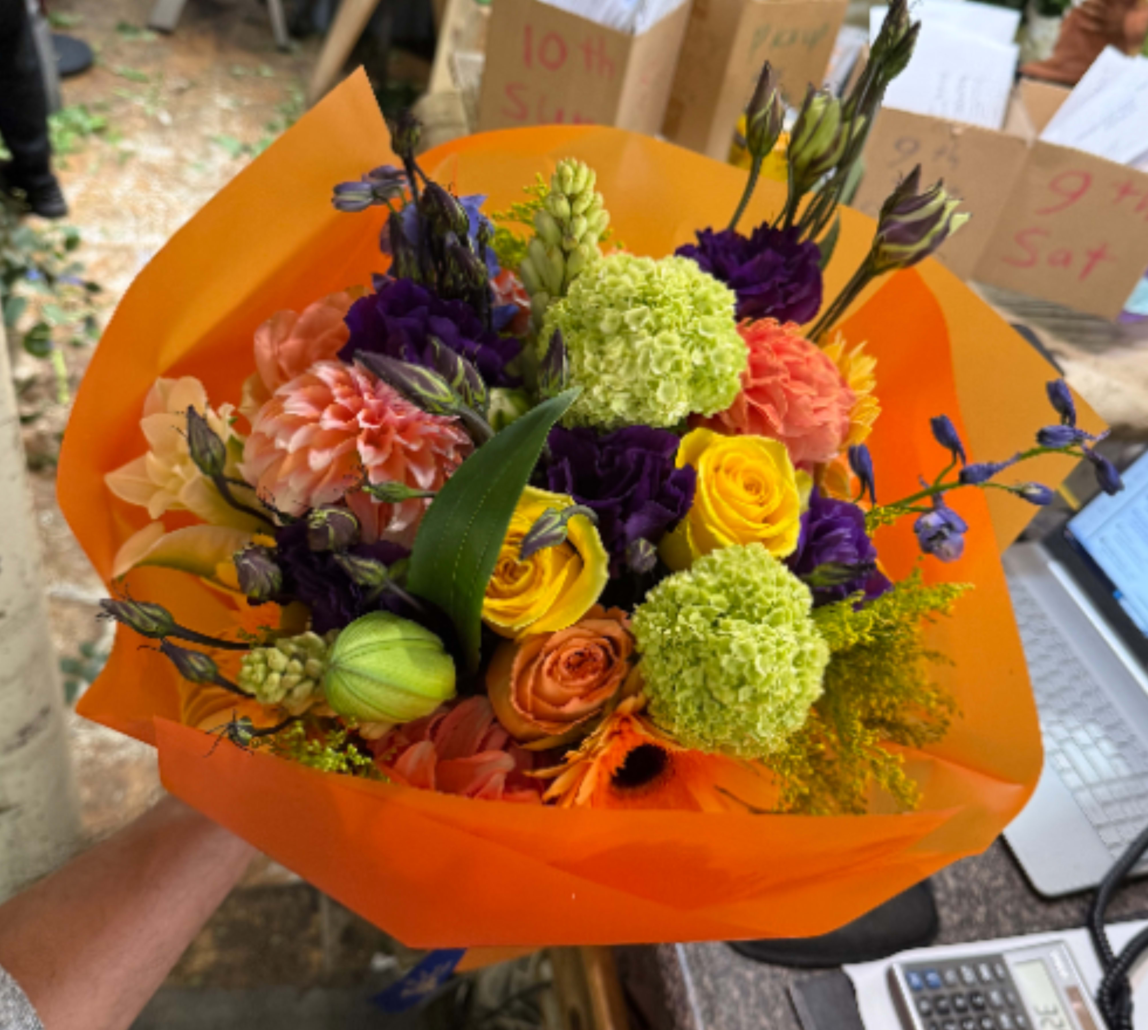 Citrus Symphony  - This bouquet is a vibrant celebration of color and texture, artfully wrapped in bold orange paper that perfectly complements its warm, cheerful palette. Featuring sunny yellow roses, peach dahlias, orange gerbera daisies, lime green viburnum, and rich purple lisianthus, it strikes a beautiful balance between balance between elegance and playfulness. The combination of bright citrus tones, soft blush hues, and deep violet accents creates a bouquet that feels energetic, modern, and full of life. 