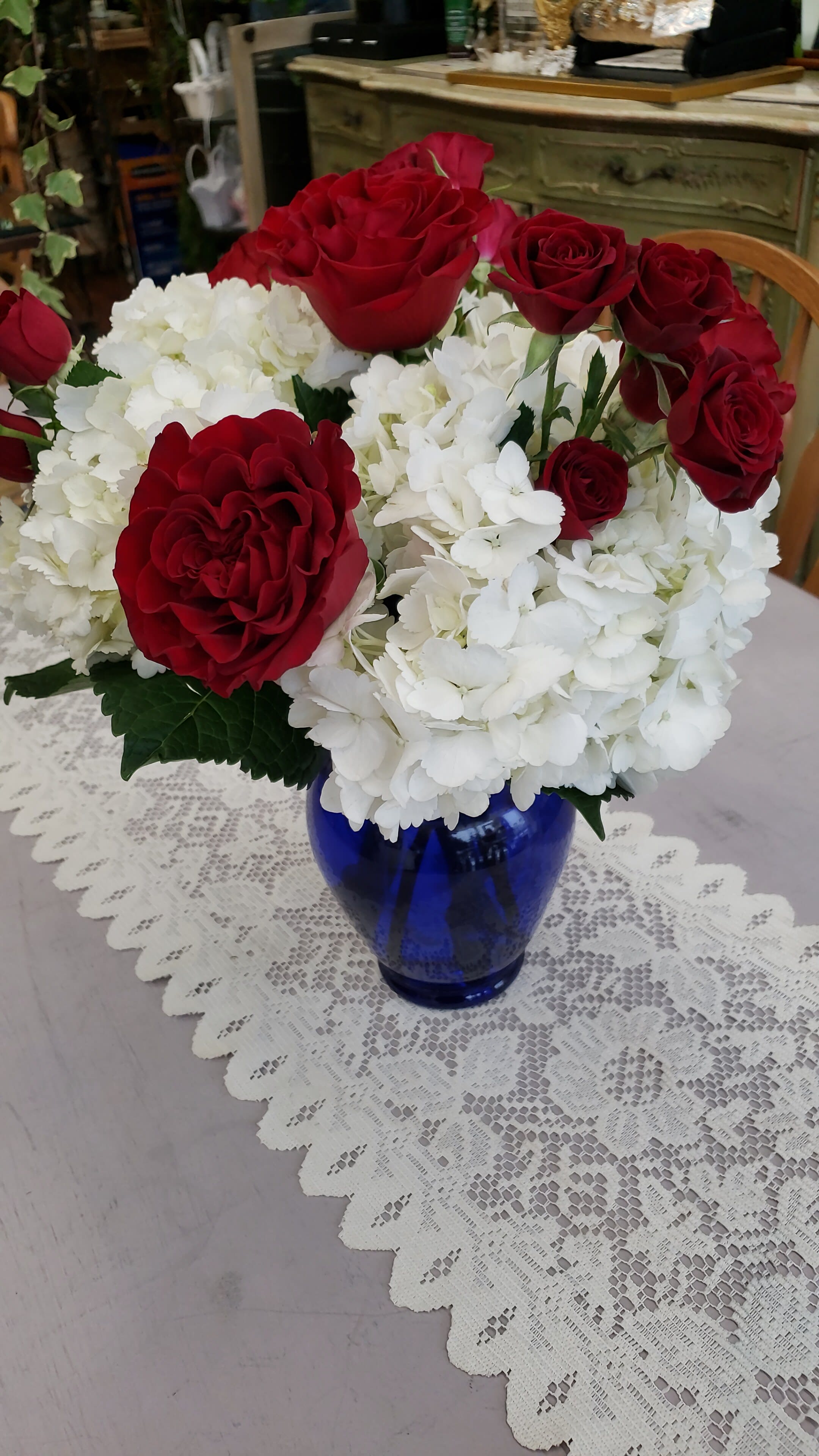 Red roses and white hydrangeas arranged in a blue glass vase on a lace table runner