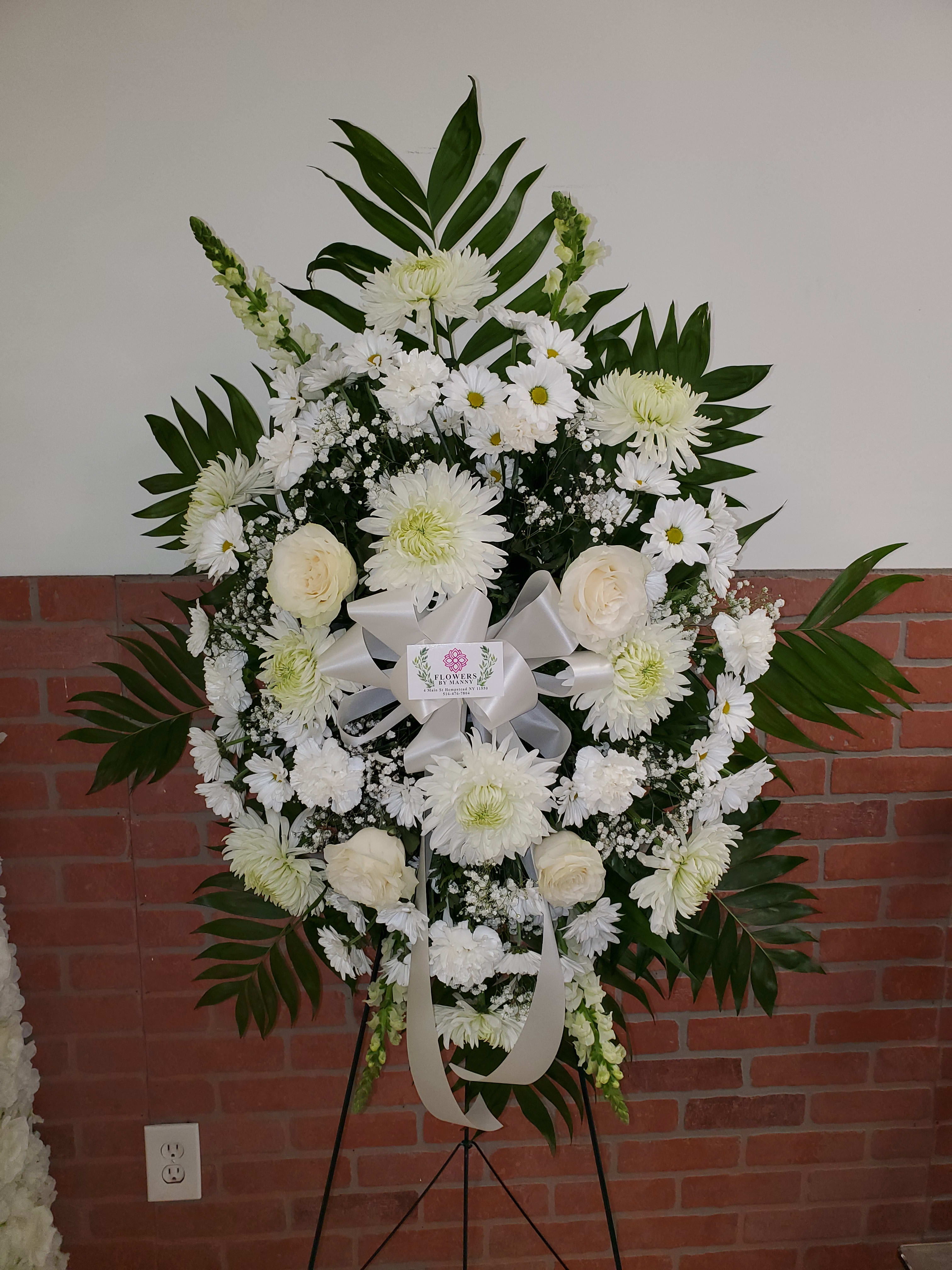 Standing white funeral spray of roses, daisies, and mums with a white ribbon on an easel.