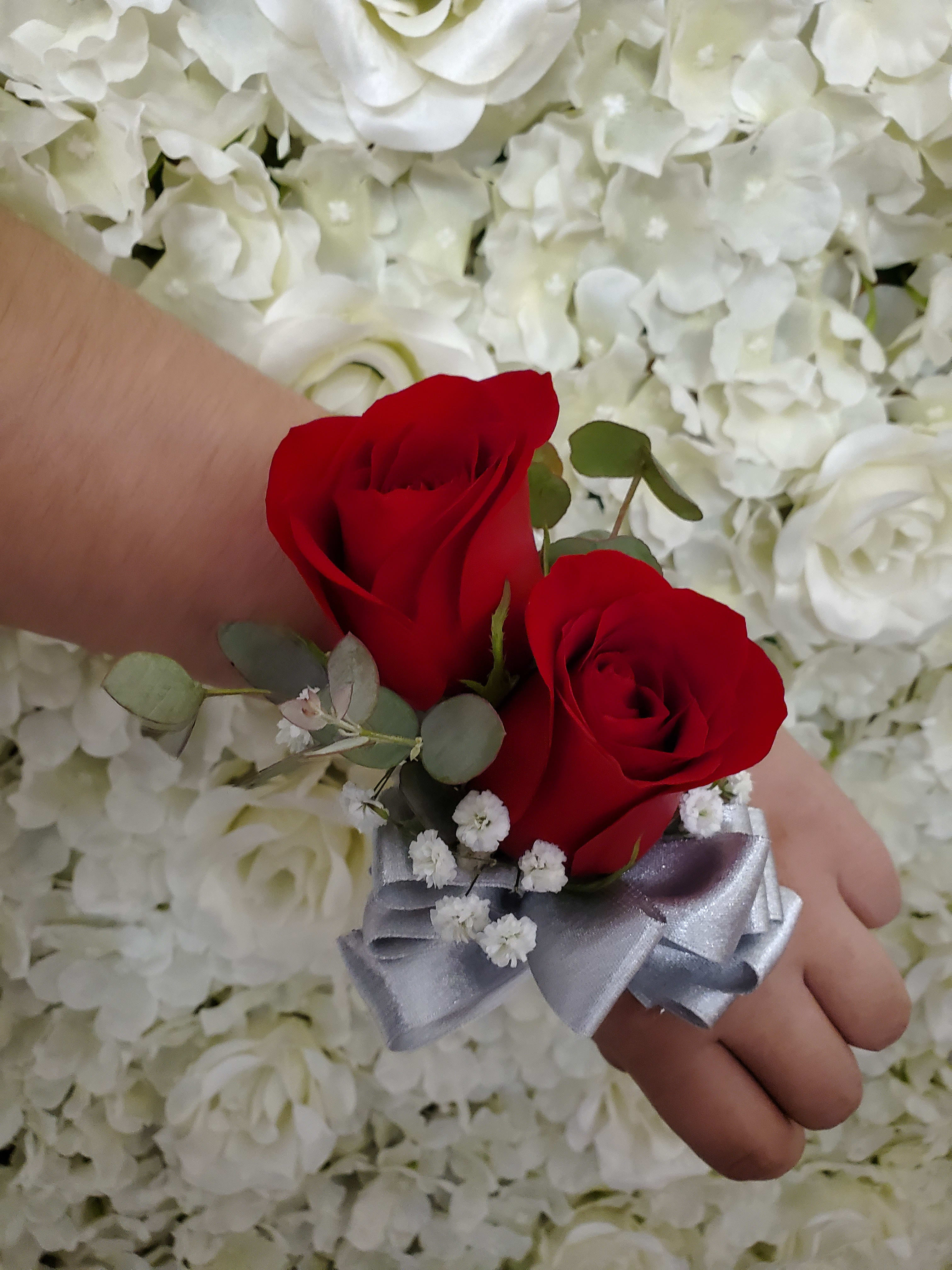 Red rose wrist corsage with silver ribbon on a hand against white flower backdrop