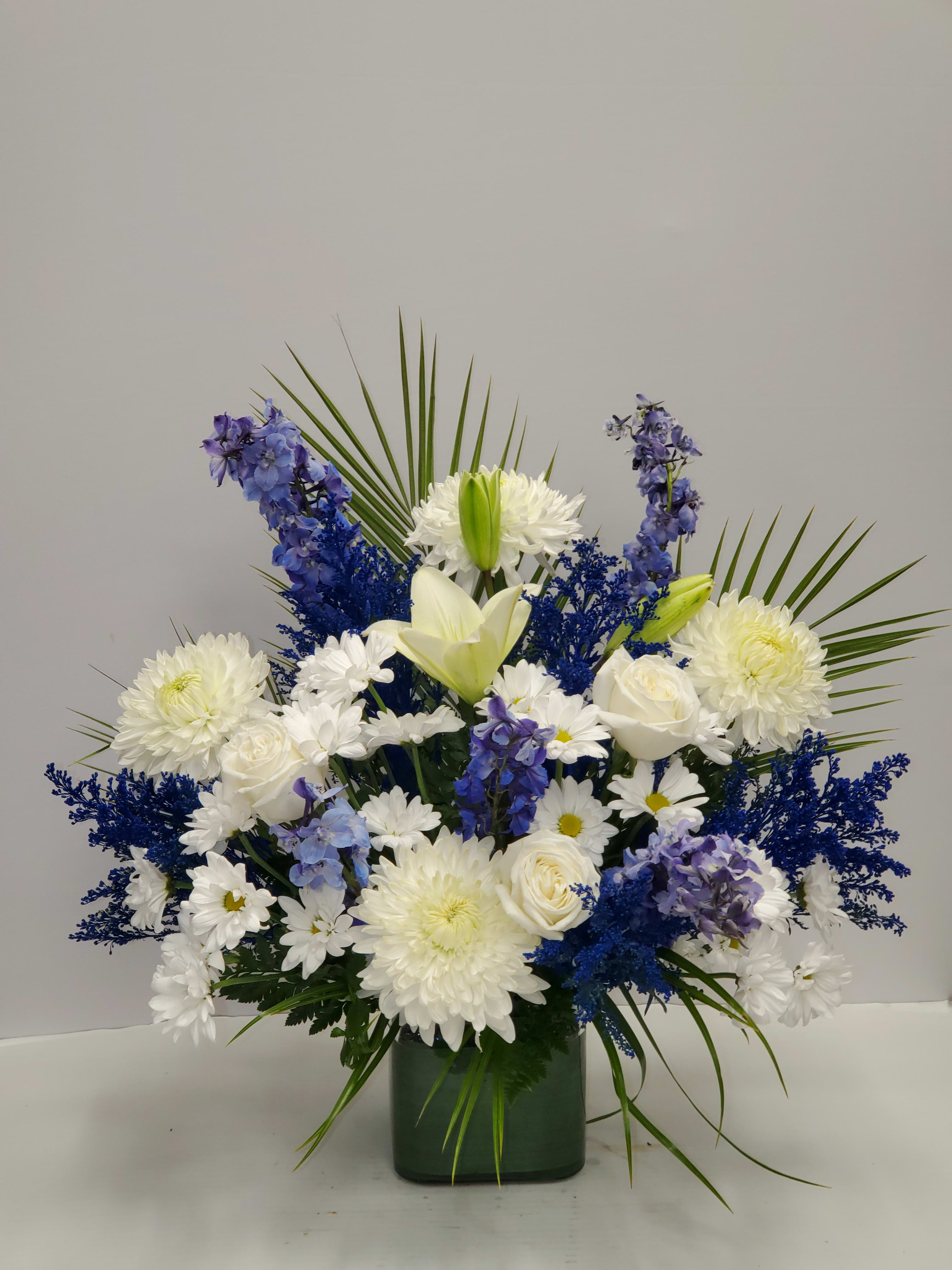 Tall arrangement of white lilies, roses, mums, and blue flowers in a green glass vase