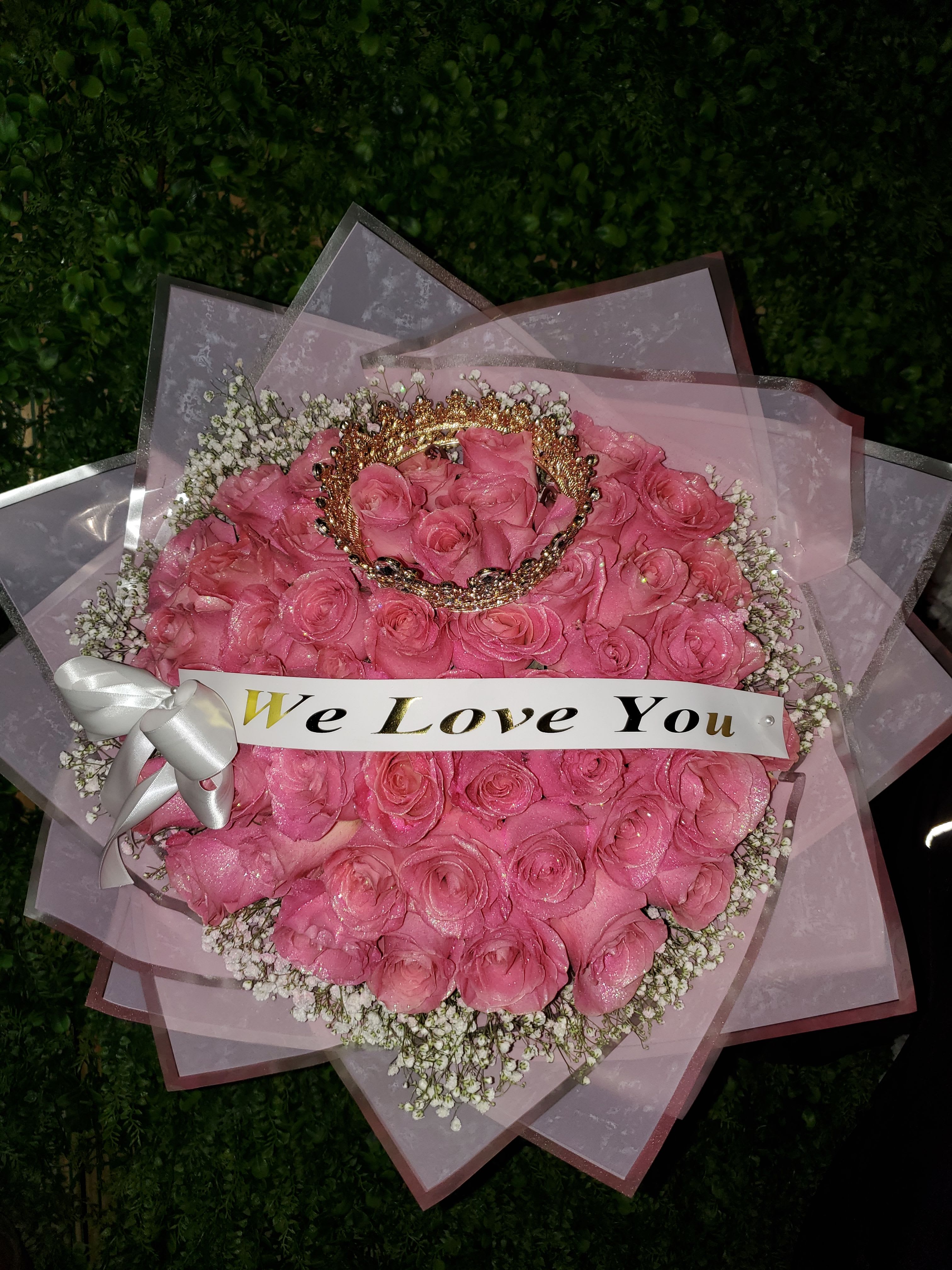 Round bouquet of pink roses with baby’s breath, crown accent, and a ‘We Love You’ banner.