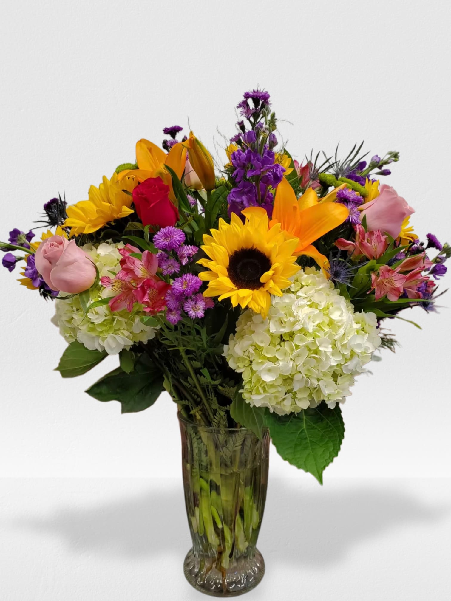 Tall mixed bouquet with sunflowers, roses, lilies, hydrangeas and purple flowers in a clear glass vase