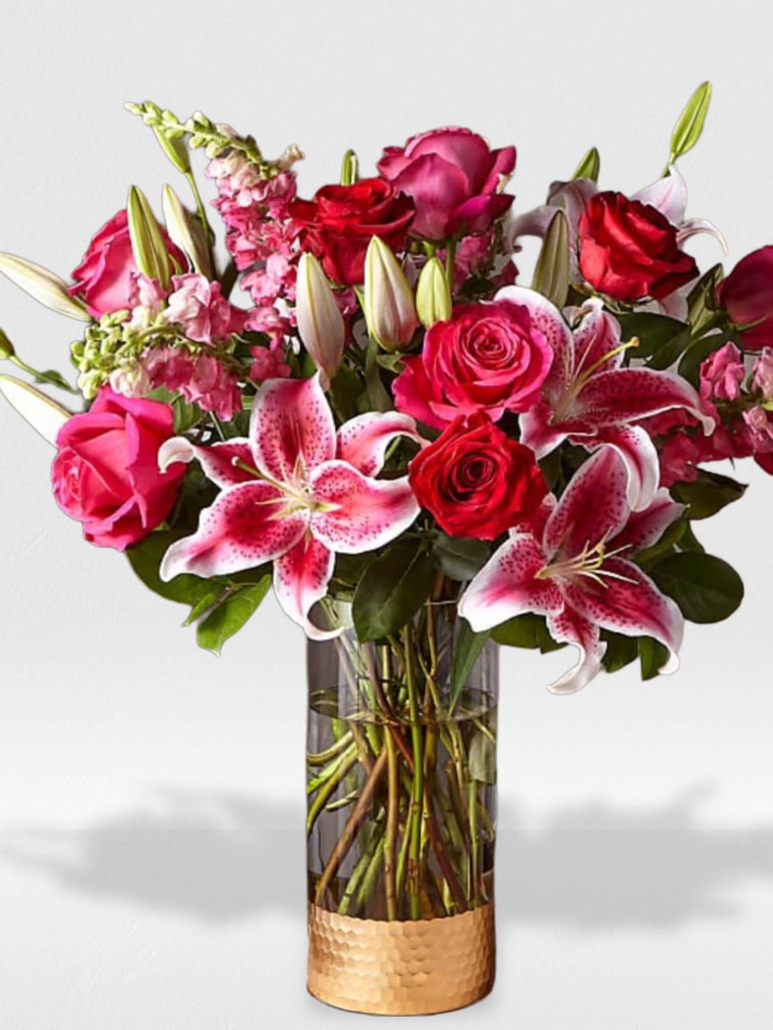 Tall arrangement of pink lilies and red roses in a clear vase with a gold band