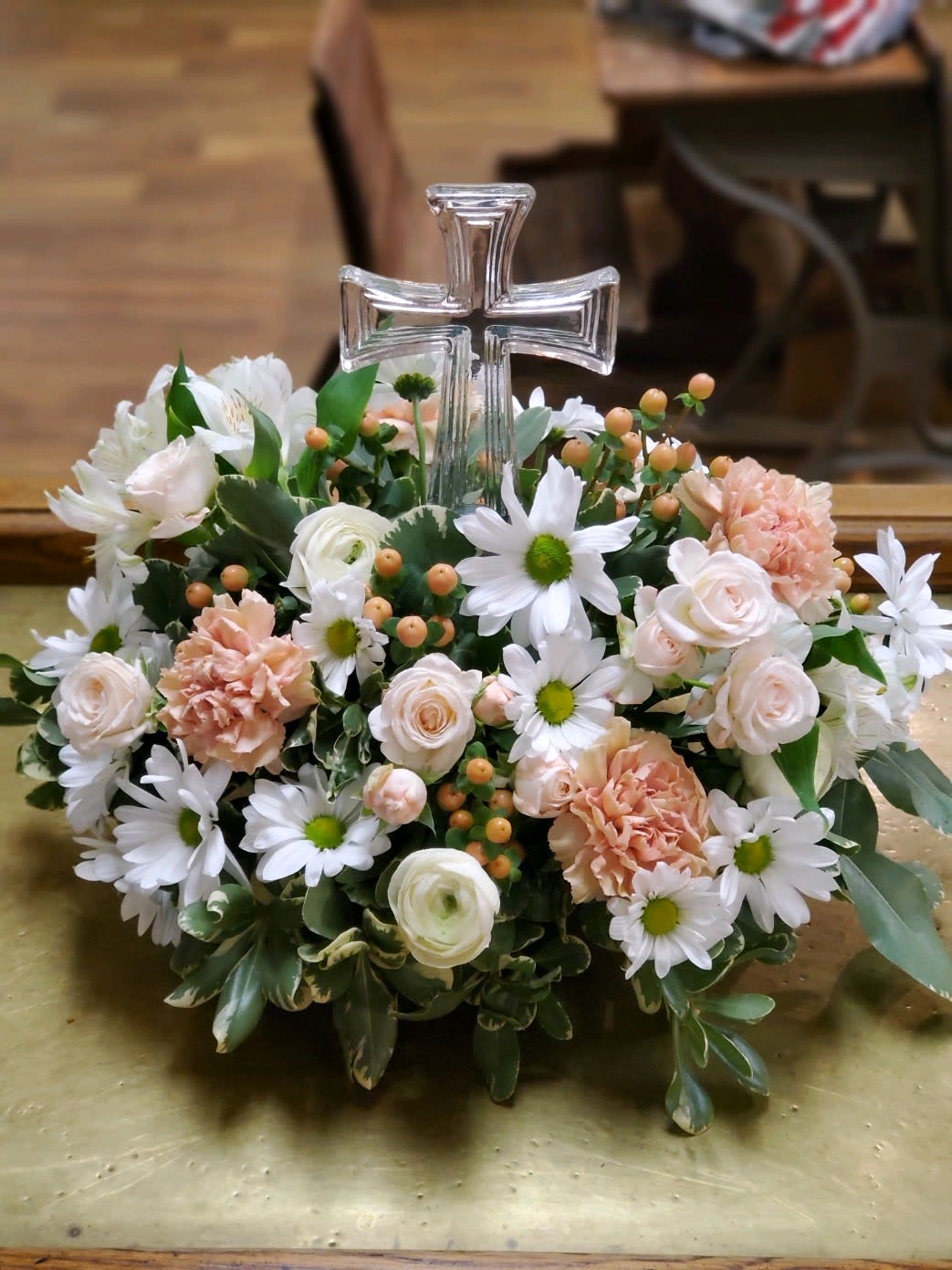 Floral arrangement with a clear cross centerpiece, white daisies, pale roses, and peach carnations