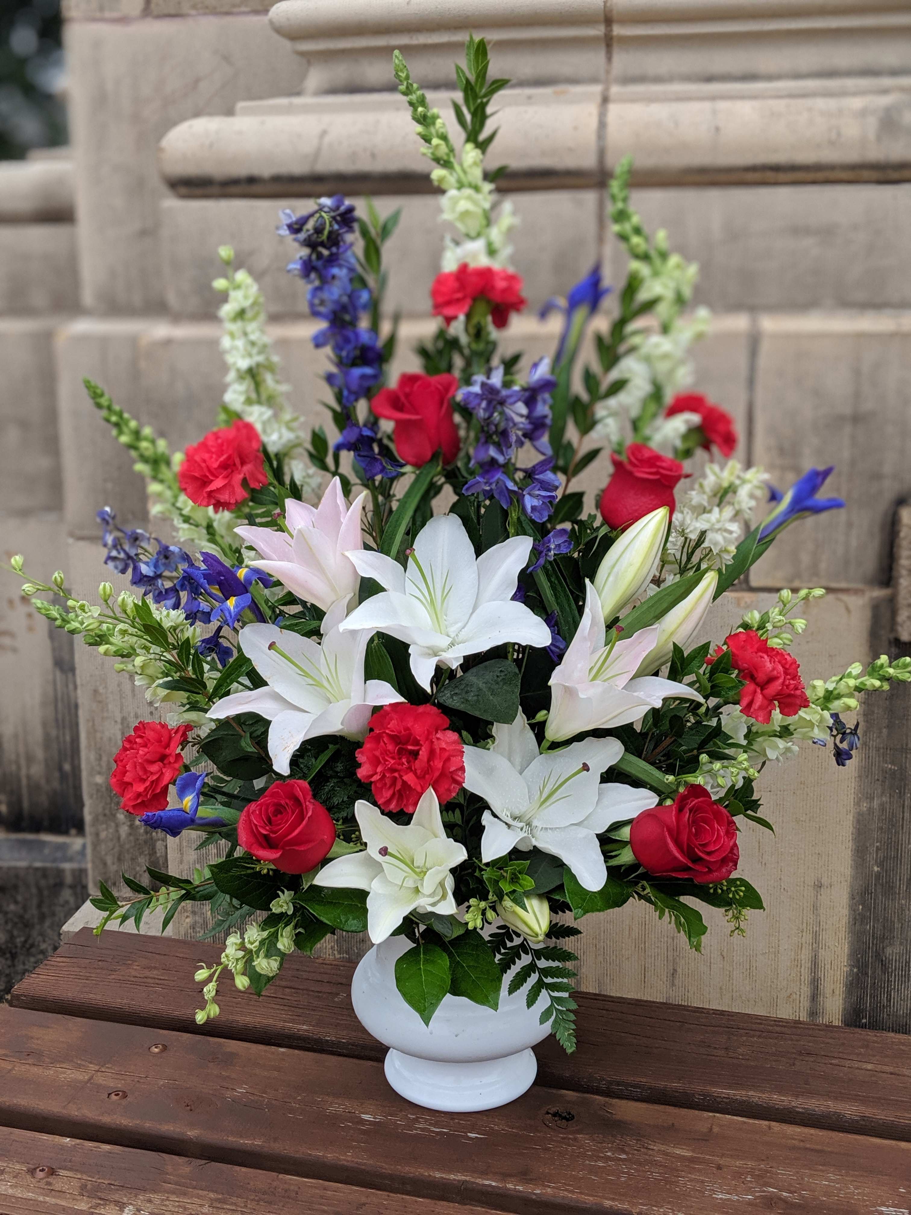 Tall red, white, and blue arrangement with lilies, roses, and carnations in a white pedestal vase