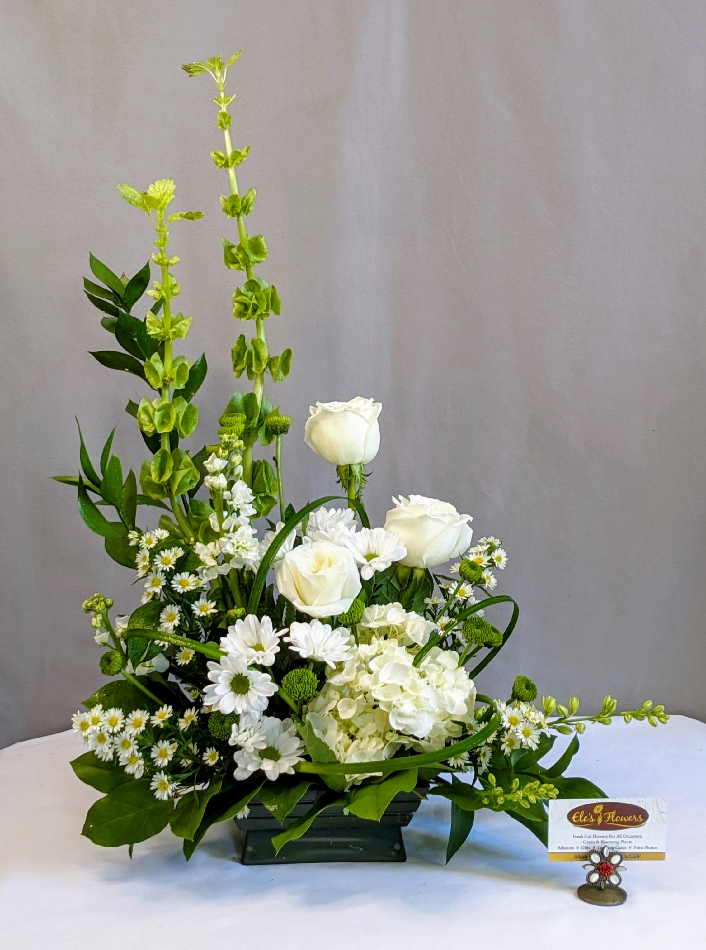 Contemporary Whites &amp; Green - Serene and elegant, Contemporary Whites &amp; Green is a calming blend of white and green blooms designed to evoke tranquility and natural beauty. This graceful arrangement features classic white roses, soft daisies, lush hydrangea, and vibrant Bells of Ireland, accented by fresh greenery and delicate accent flowers. Arranged in a modern container, it offers a refined yet organic feel—perfect for expressing sympathy, offering comfort, or bringing a sense of calm to any space. Understated and timeless, this design speaks gently when words are hard to find.