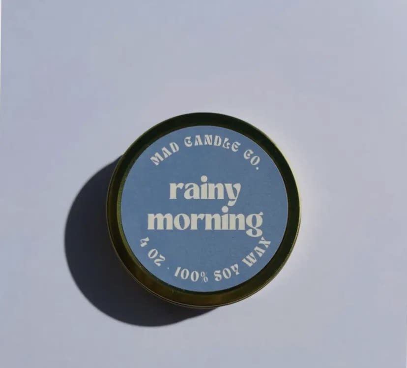 Round candle tin labeled "rainy morning" on a plain surface
