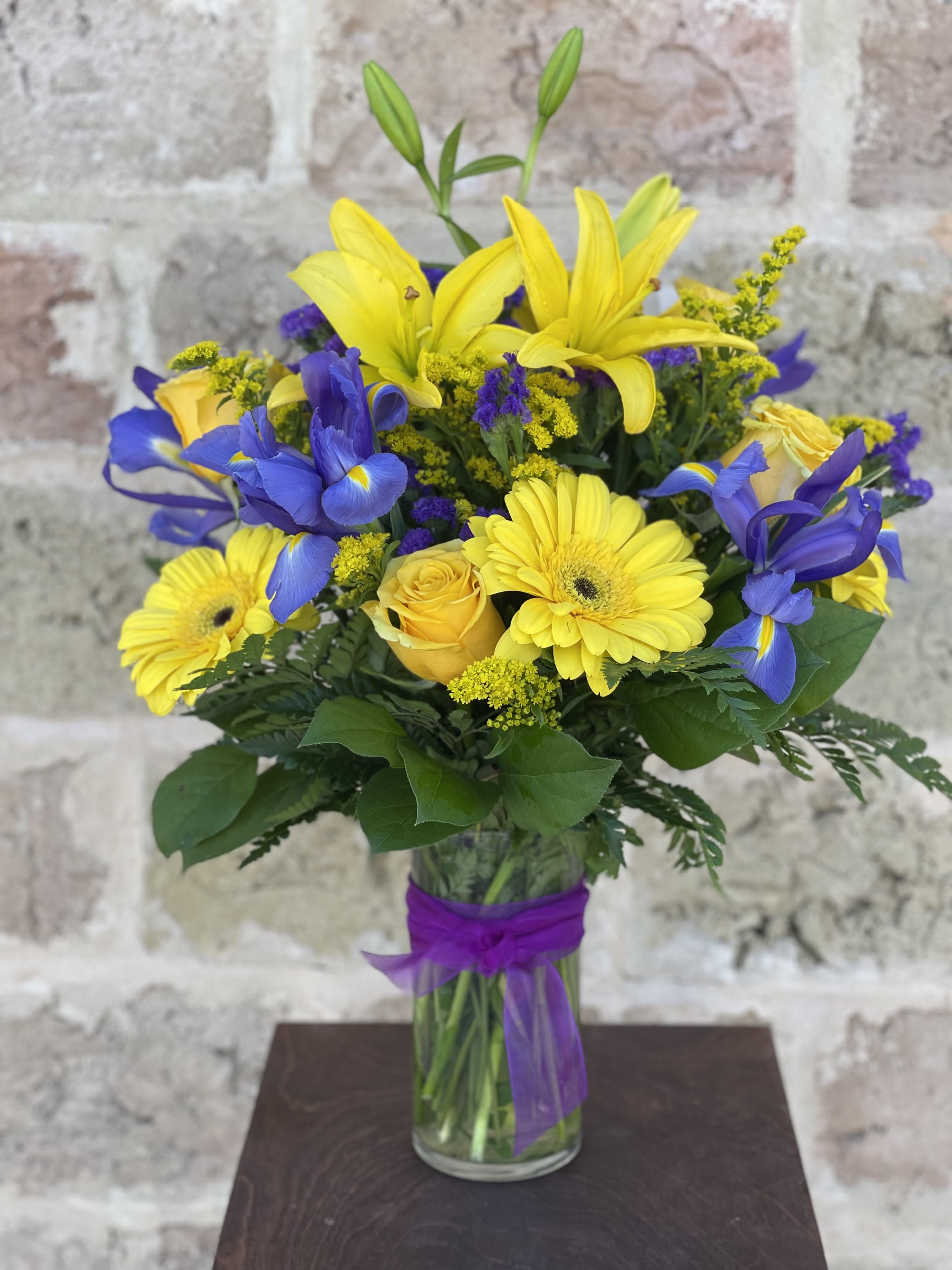 Yellow lilies, blue irises, and yellow daisies in a glass vase