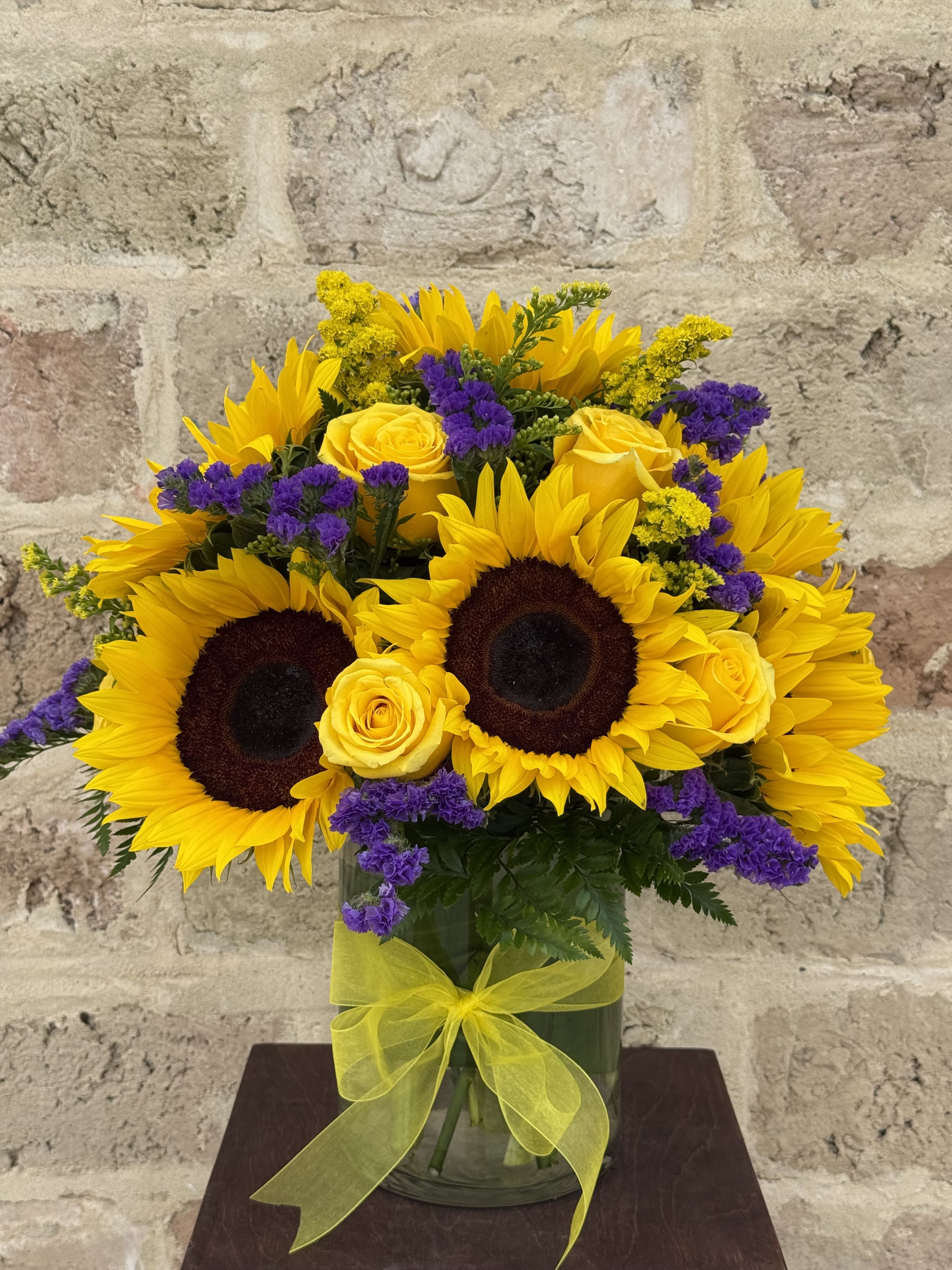 Sunflowers, yellow roses, and purple filler flowers in a glass vase with a yellow ribbon.