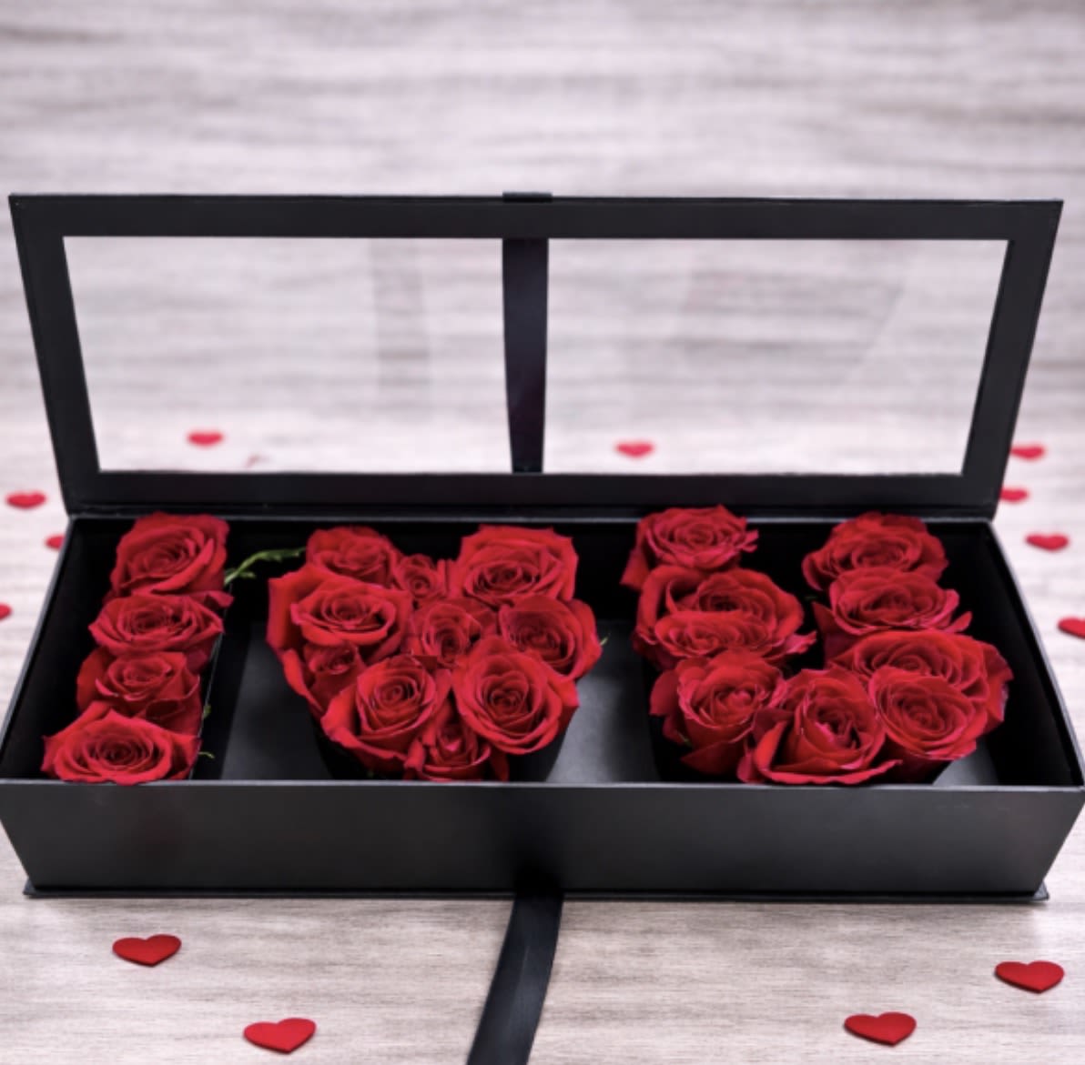 Red roses arranged in a black rectangular box with clear lid, surrounded by small red heart accents