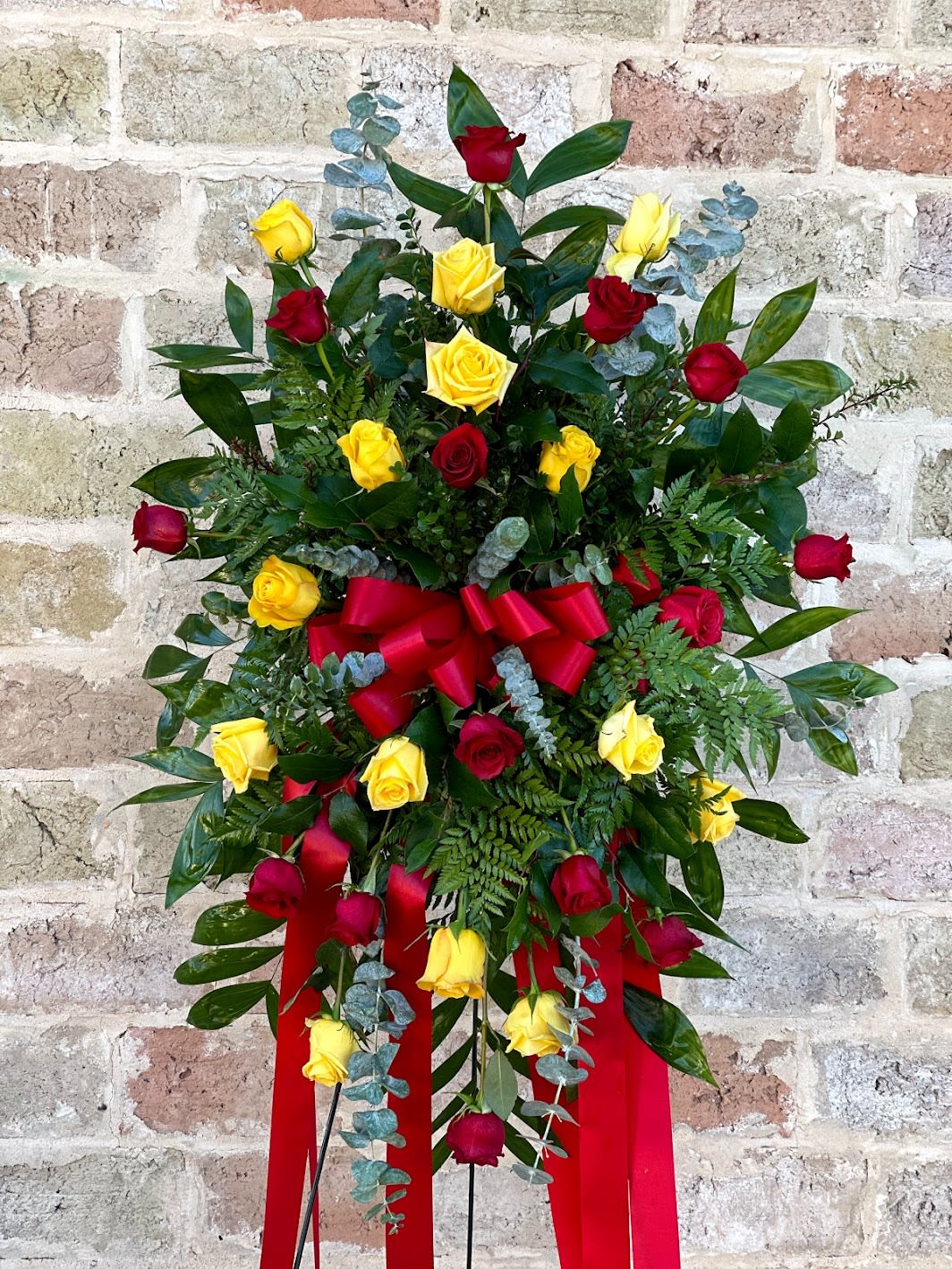 YELLOW AND RED ROSES STANDING SPRAY - A tall standing spray of red and yellow roses makes a bold, heartfelt statement for a sympathy or funeral service. The combination of crimson and sunny blooms reads as both reverent and uplifting-an arrangement that helps the person you're honoring feel truly remembered and the family feel comforted in a time of loss.  Clusters of long-stem red and yellow roses are nestled into dense greenery and silver-green eucalyptus for rich texture and contrast, finished with crisp white accents. A large red satin bow with long ribbon tails is tied near the center for a formal, traditional presentation, and the arrangement is mounted on a standing easel for easy display at a service. The proportions lend vertical presence while keeping the overall look balanced and dignified.  Ideal for honoring a loved one or sending condolences that feel both personal and graceful, this standing spray conveys love, respect, and celebration of life. Standard care for display: keep away from direct heat and replace water source per funeral home guidelines; sizing and delivery details are available at checkout to ensure it fits the service space.