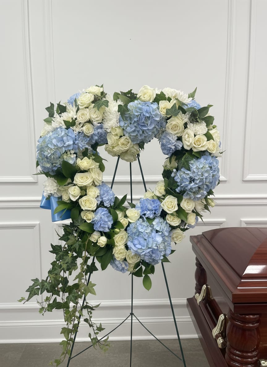 Blue-ming Heart  - A  heart-shaped sympathy wreath that reads as a calming, dignified tribute-perfect for a funeral or memorial service. This heart-shaped arrangement on a metal easel stand combines pale blue hydrangeas with pure white mini roses and chrysanthemums to create a serene blue-and-white palette that comforts and honors the person you're remembering.  The wreath arrives ready to display on the metal stand; for best appearance, keep it sheltered from direct sun and extreme heat until presentation.