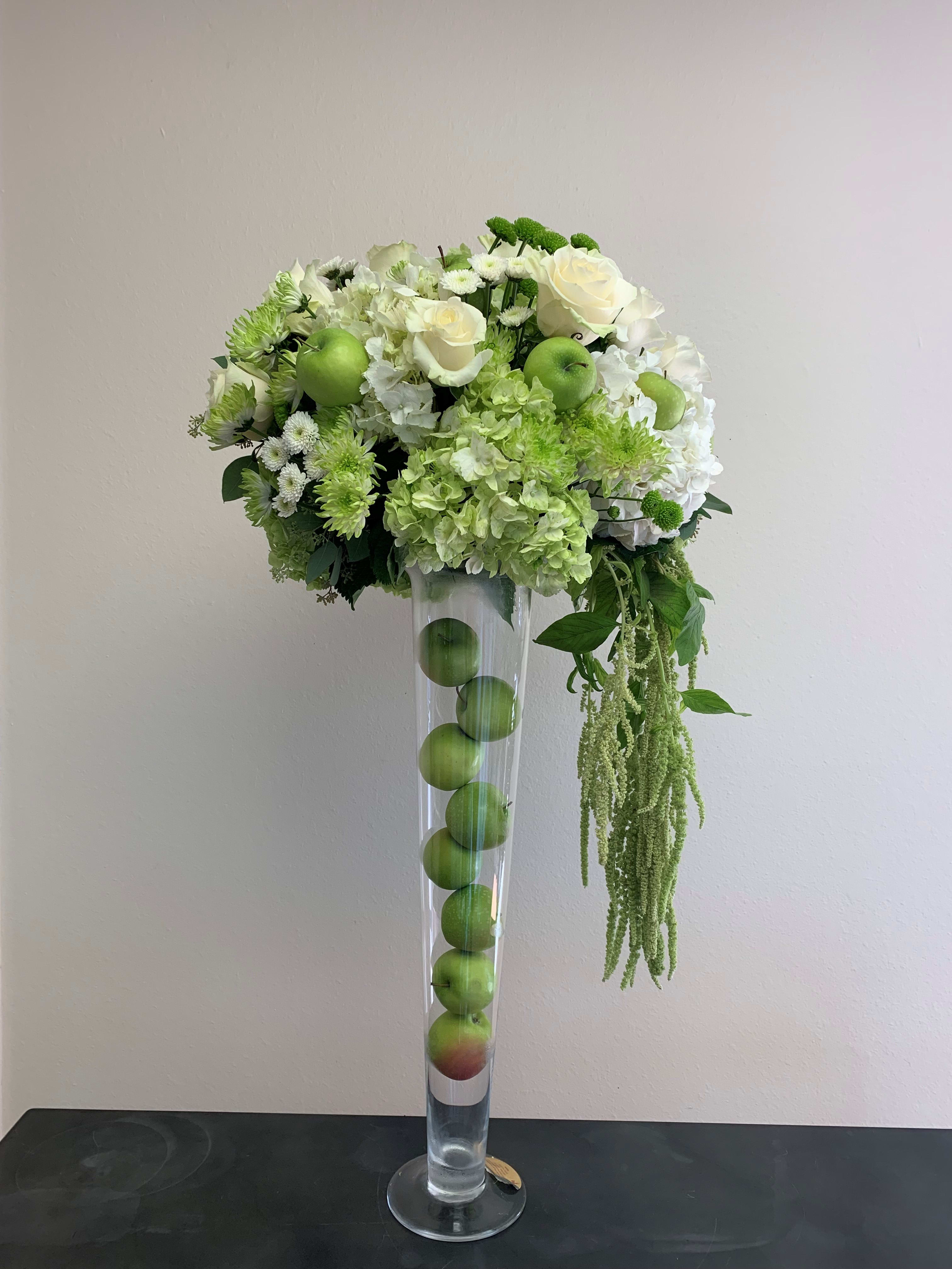 White and green floral arrangement in a tall clear vase with apples inside