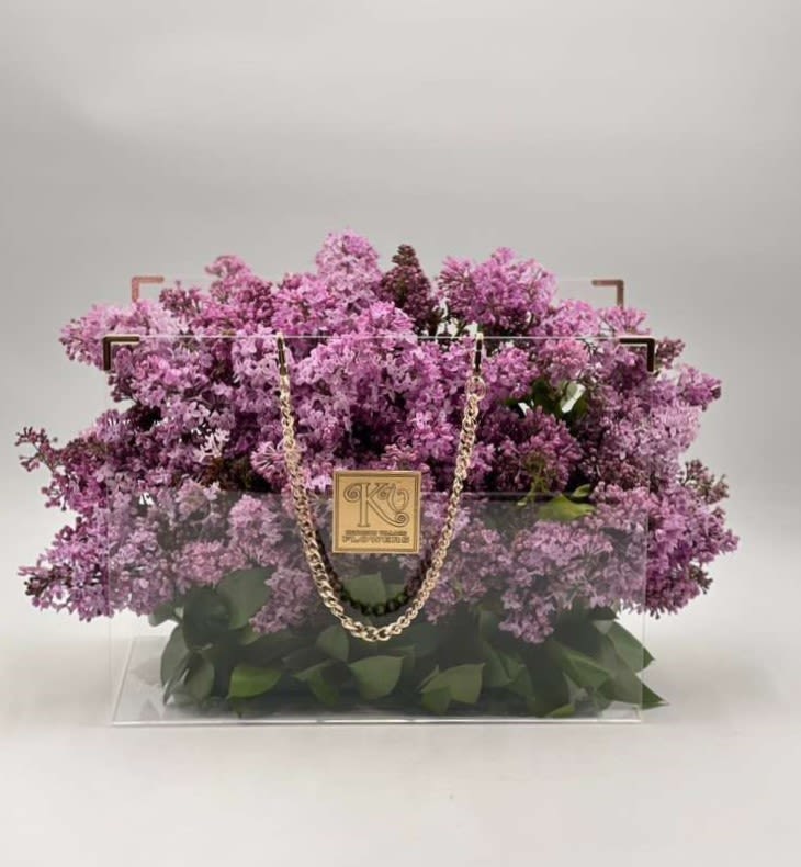 Lilac Luxe Handbag - Elegant, fragrant, and full of spring charm, this stunning arrangement showcases lush clusters of fresh lilac blooms beautifully arranged inside a modern clear handbag-style container accented with a delicate gold chain. The soft lavender blossoms create a cloud-like, romantic texture while the fresh greenery at the base adds natural depth and balance. Designed to capture the beauty of spring in a sophisticated presentation, this arrangement makes a perfect gift for birthdays, celebrations, or anyone who loves the timeless fragrance and elegance of lilacs.