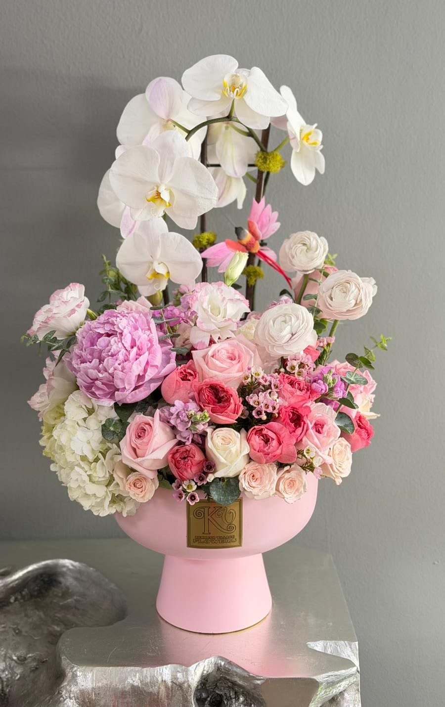 Orchid Elegance - An exquisite arrangement featuring graceful white orchids, lush pink peonies, premium garden roses, ranunculus, hydrangea, and delicate seasonal blooms, artfully designed in a modern blush pedestal vase. A timeless expression of elegance, love, and sophistication.