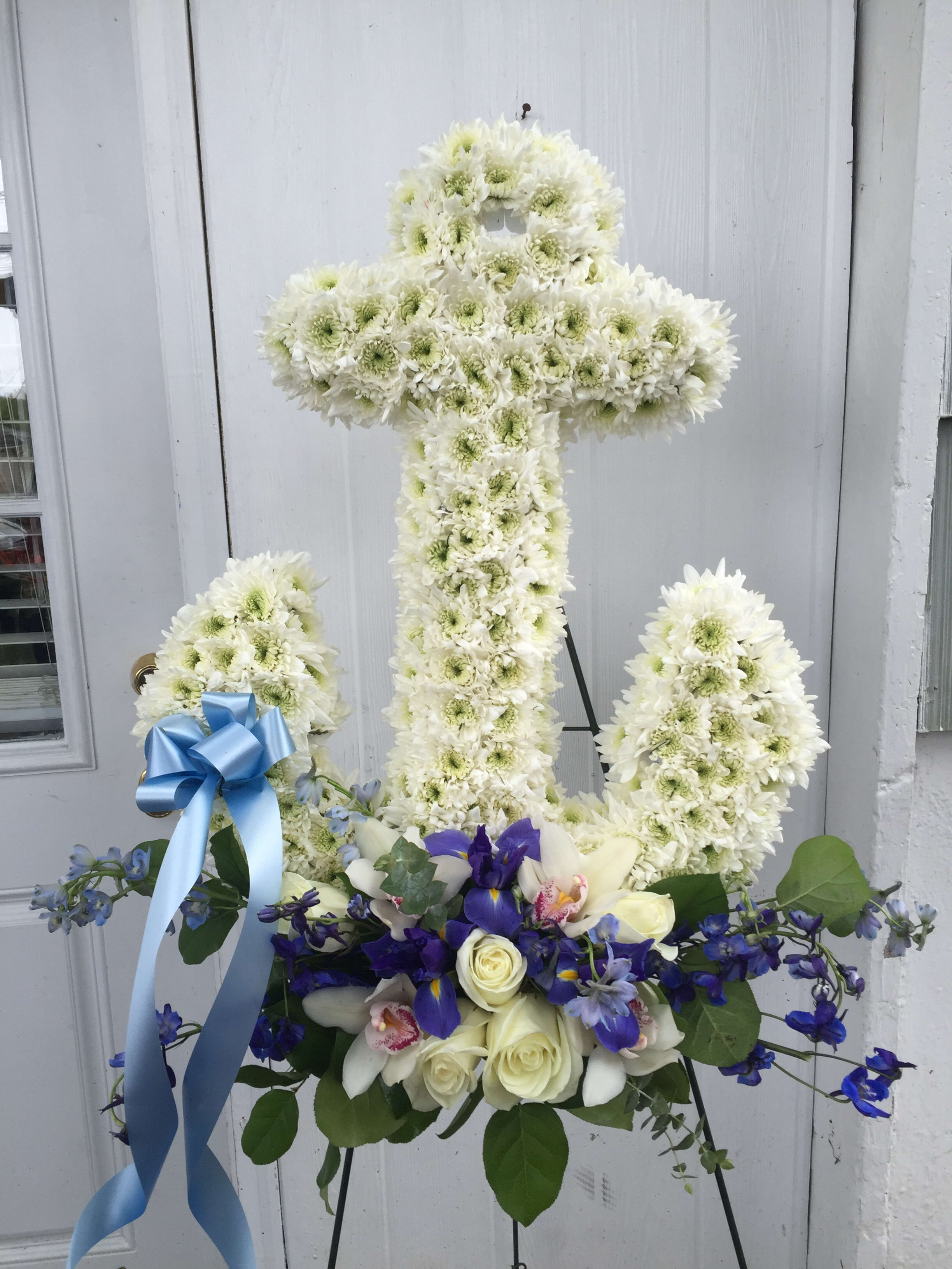 White floral cross on an easel with blue ribbon and purple flowers at the base