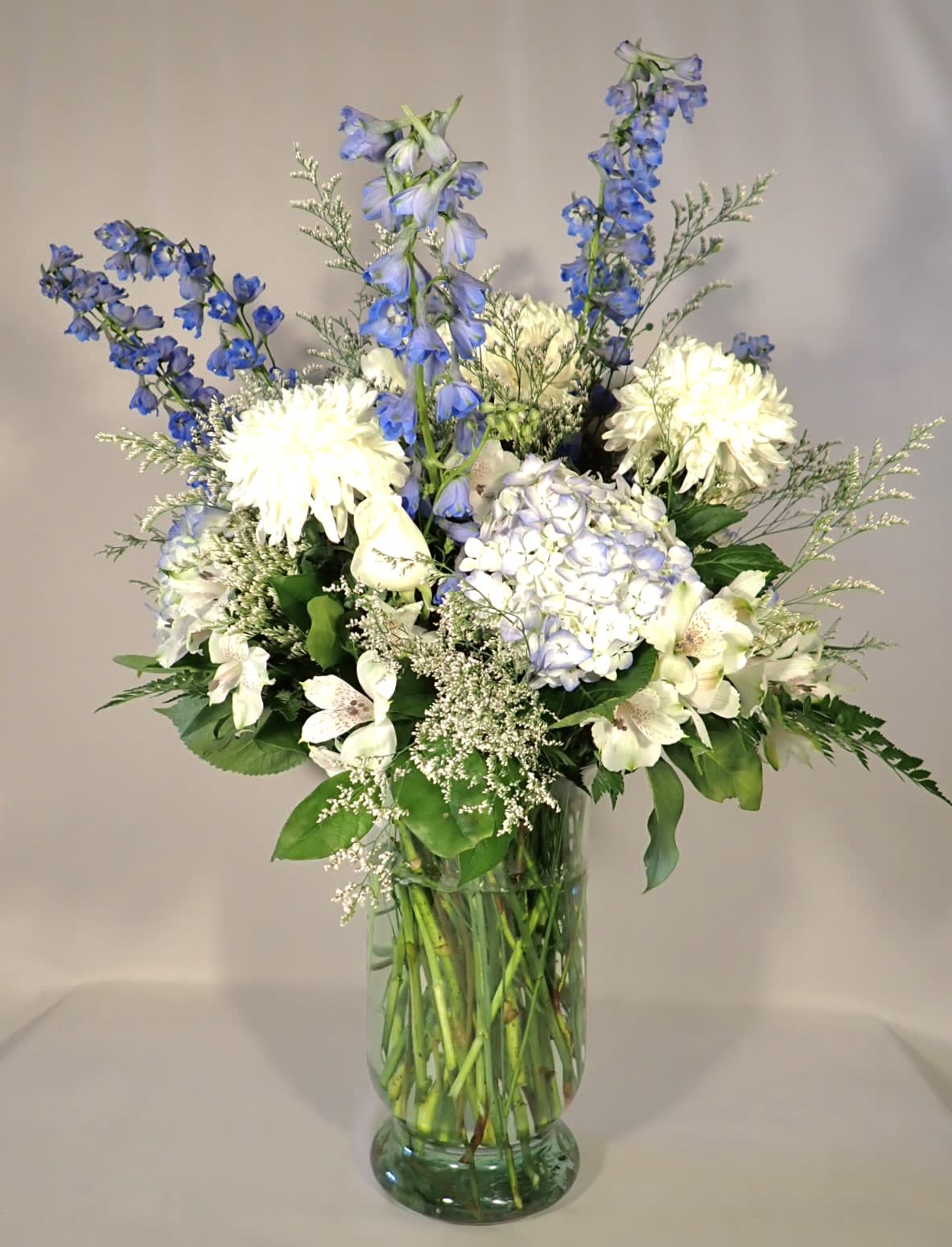 Sky Delight - This beautiful arrangement of blue Hydrangeas, white Roses, white Magnums, white Alstroemeria, white Limonium, blue Delphinium, Salal and Leather Leaf in a large glass vase picturesque of the summer day sky. 30"H 22" W 