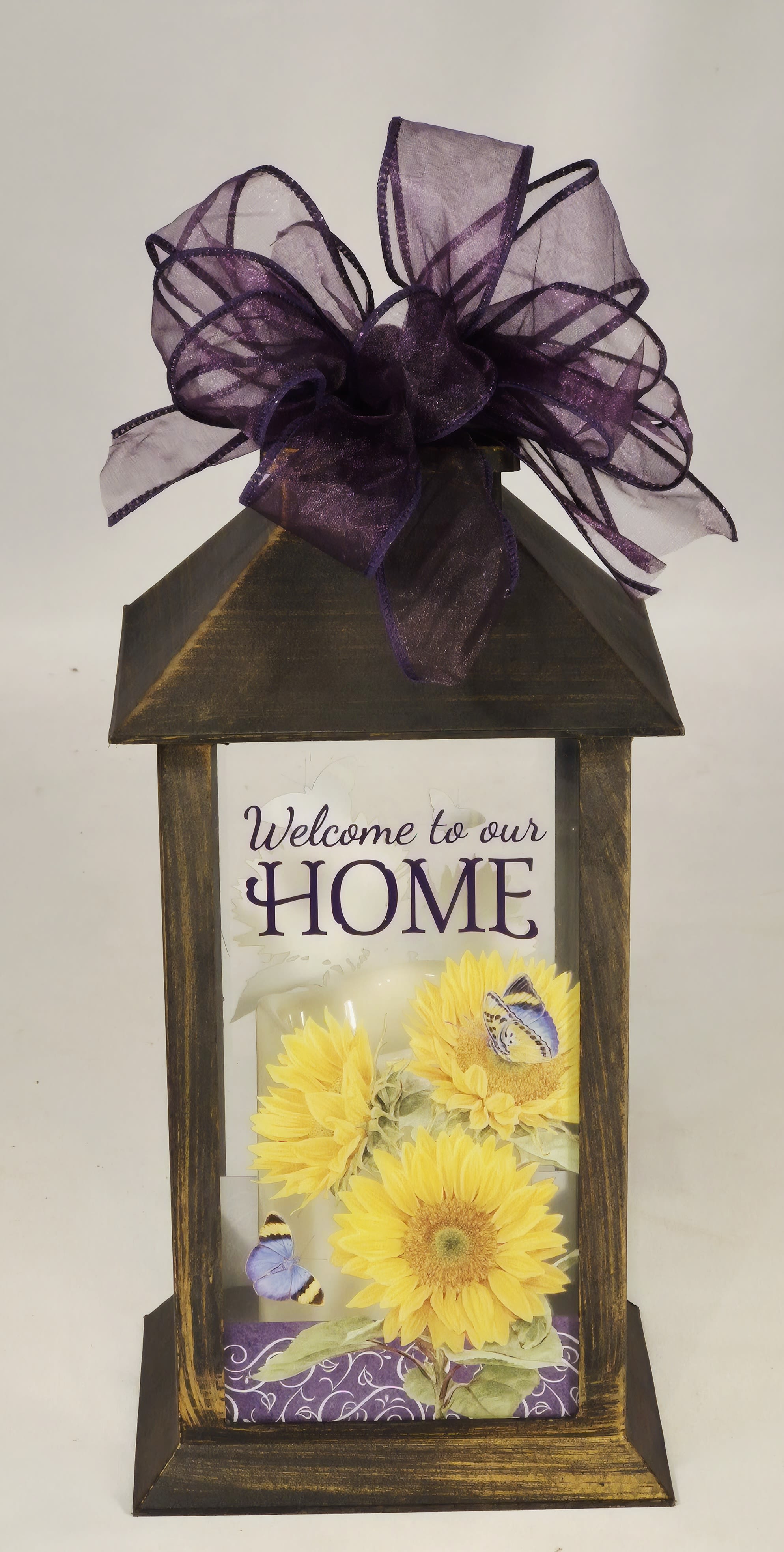 Welcome to Our Home Lighted Lantern - Bright yellow sunflowers take center stage in this Welcome to Our Home Sweet Sunflowers Lighted Lantern, a cheerful housewarming flower arrangement designed to greet guests and make someone feel instantly at home. The vertical, rustic display pairs sunny blooms with clusters of purple flowers and broad speckled greenery, creating a bold contrast that reads fresh and friendly-ideal for summer decor, a thoughtful thank-you gift, or a spontaneous "just because" surprise.  The arrangement is set inside a dark wooden lantern-style frame with a printed "Welcome to our HOME" sign on UV-printed glass, and finished with a decorative bow. The lantern includes a flameless LED candle with a built-in timer (6 hours on / 18 hours off) and comes ready to hang or sit on an entry table or porch;   W: 5.5" H: 13" D: 5.5