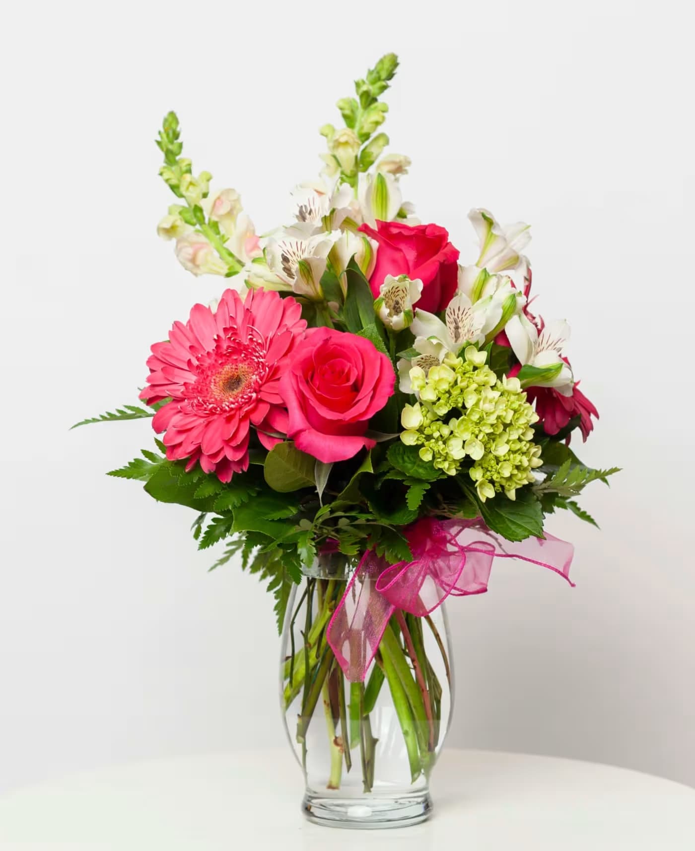 Pink gerbera daisies and roses in a clear glass vase with a pink ribbon