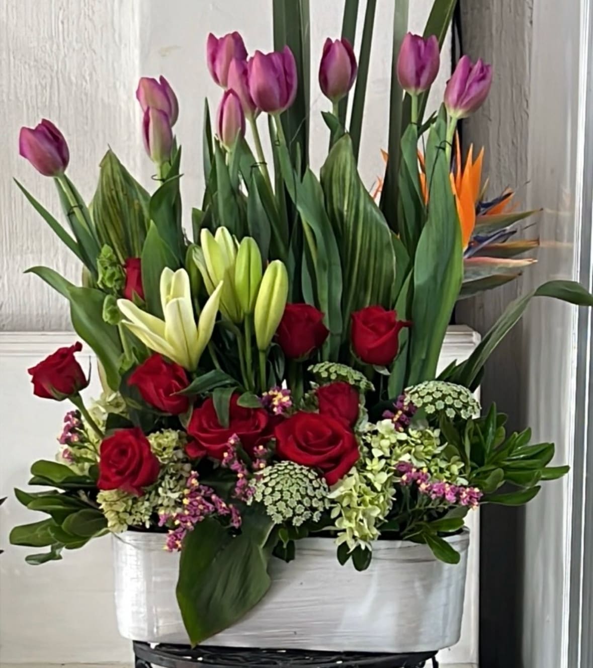 Tall floral arrangement with red roses, purple tulips, and white lilies in a white container