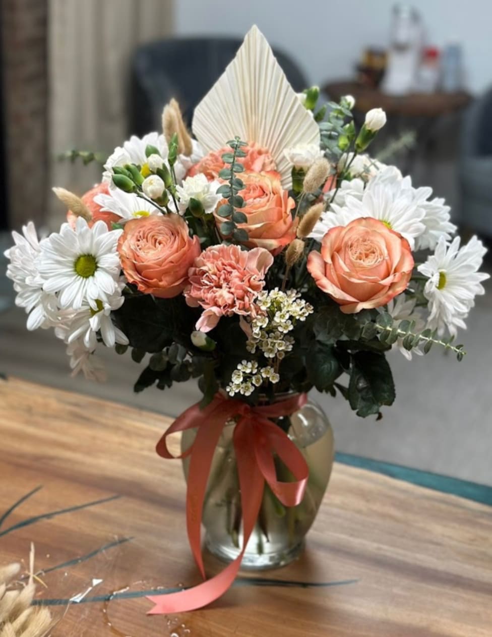 Peach roses and white daisies arranged in a glass vase with a red ribbon