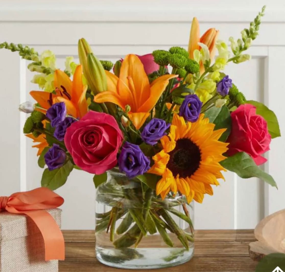 Bright mixed bouquet in a clear glass vase with roses, lilies, sunflowers, and purple blooms