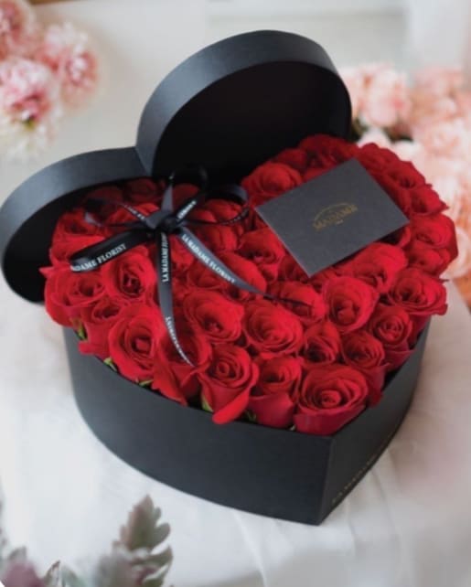 Red roses arranged in a black heart-shaped box with a ribbon and card